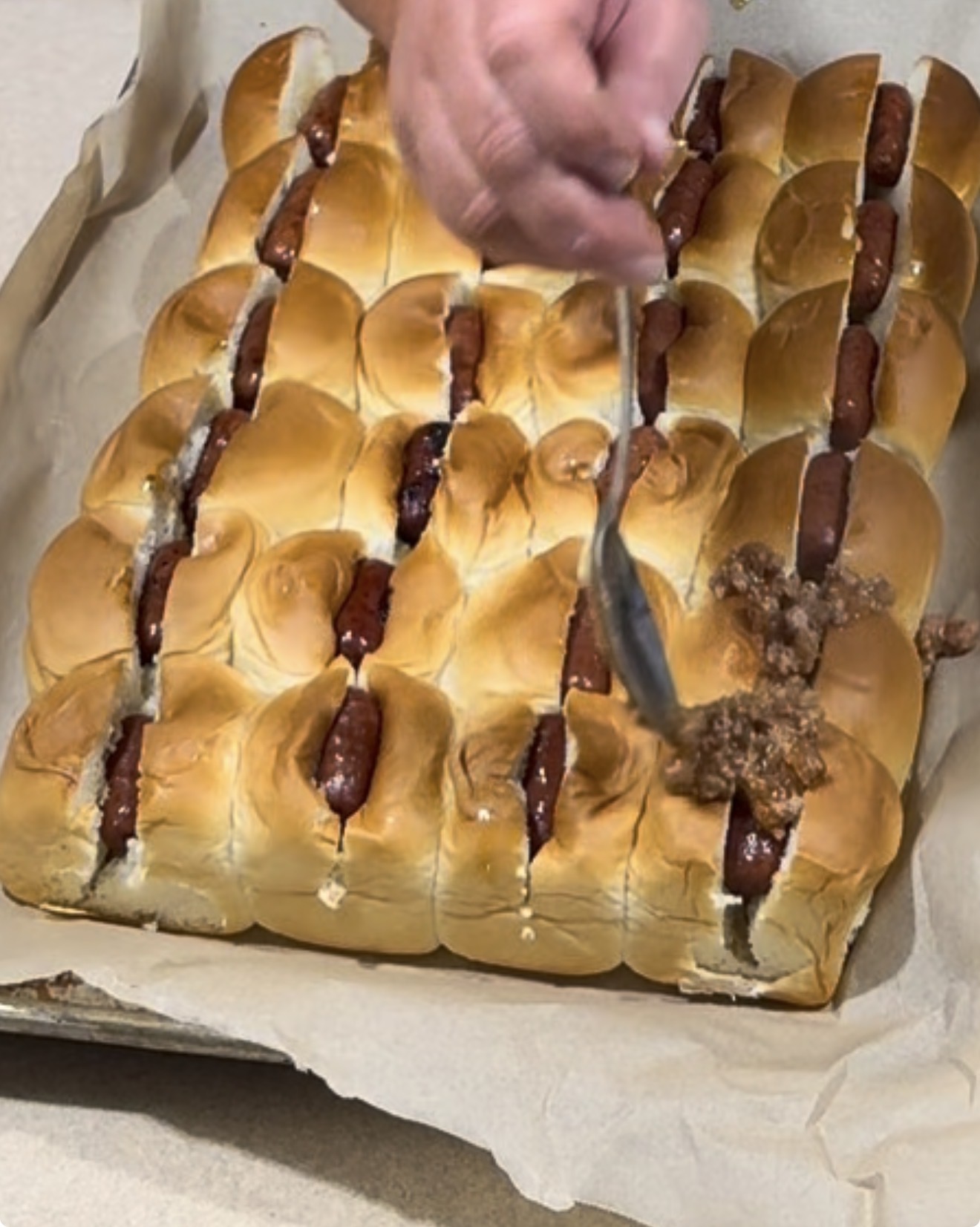 A hand uses a spoon to add ground beef topping onto rows of hot dogs in split buns, arranged closely together on a parchment-lined baking sheet.