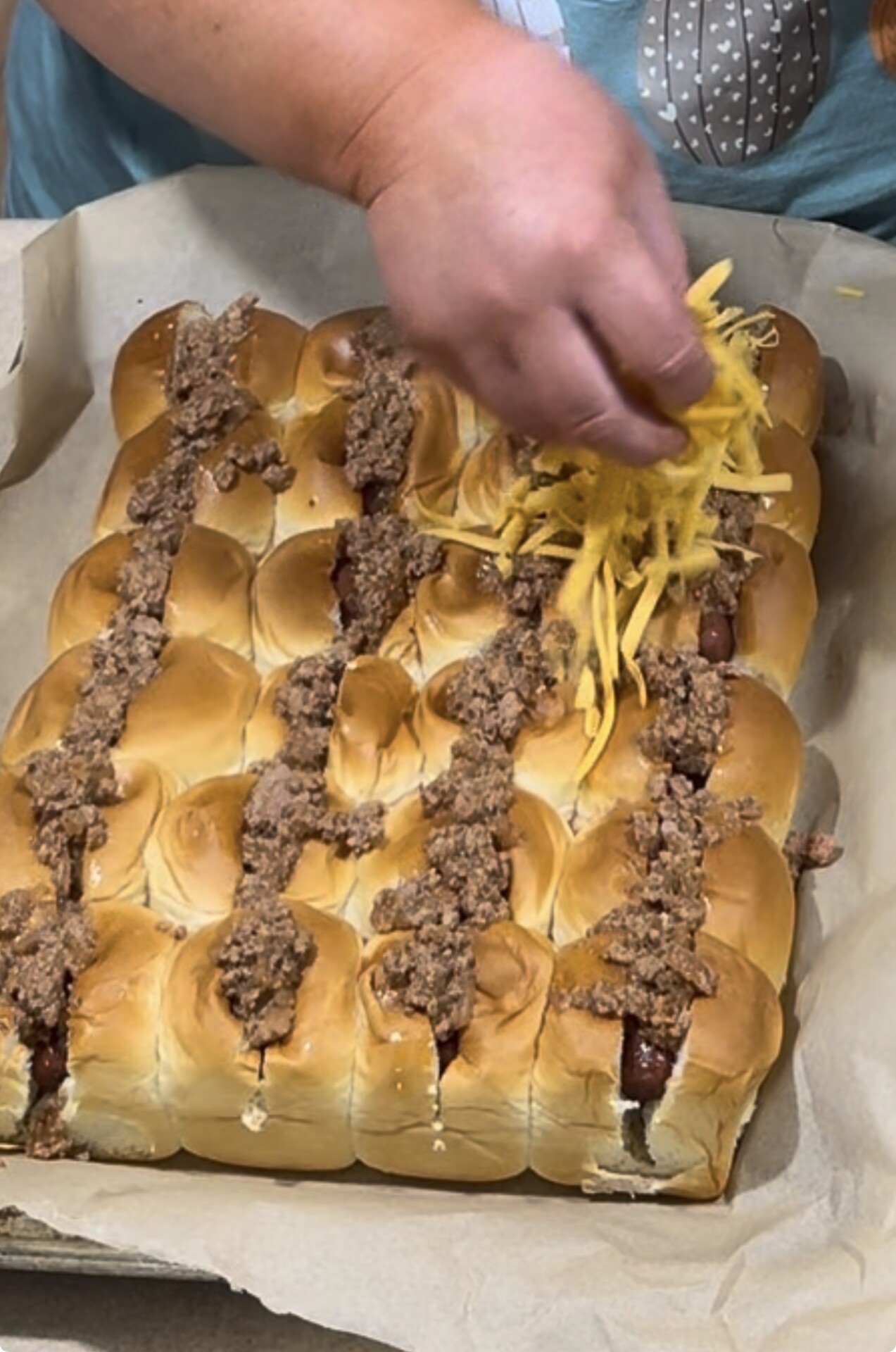 A hand sprinkles shredded cheese over a tray of small hot dogs in buns topped with cooked ground beef, all arranged closely on parchment paper.