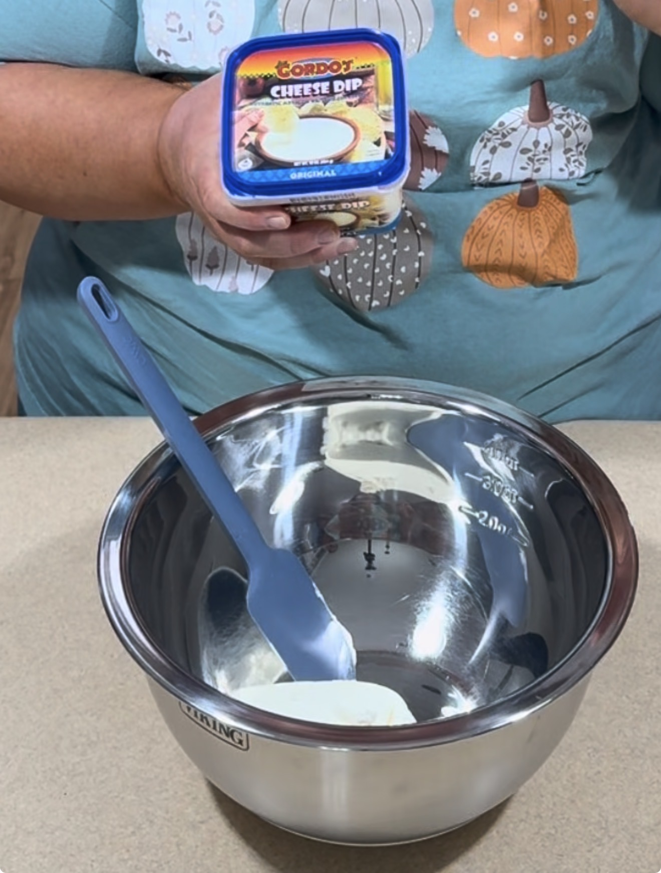 A person holds a tub of cheese dip above a metal mixing bowl on a countertop, with a blue spatula resting inside the bowl. The person is wearing a teal shirt with pumpkin designs.