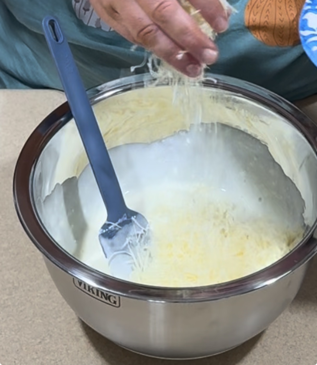 A person sprinkles shredded cheese into a stainless steel mixing bowl with a spatula resting inside, preparing a mixture on a kitchen counter.