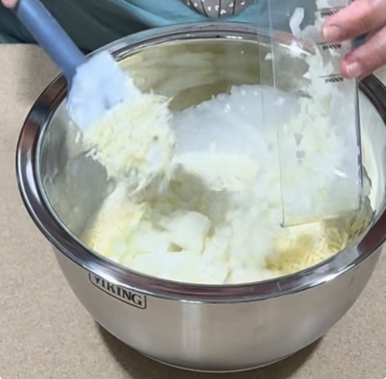 A person uses a spatula to mix shredded cheese, onion, and other ingredients in a large stainless steel mixing bowl on a countertop.