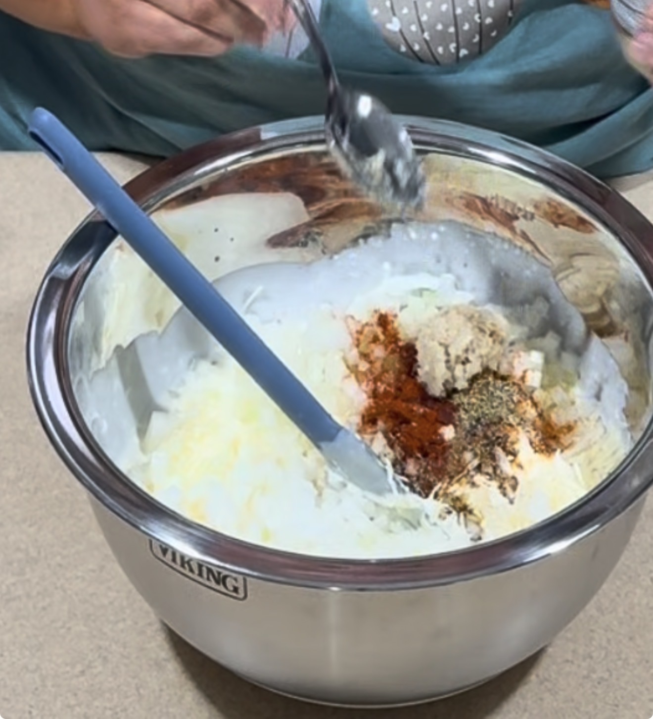 A mixing bowl with shredded potatoes, spices, and grated cheese. A blue spatula rests inside, and a person holds a spoon above the bowl, preparing to mix the ingredients.