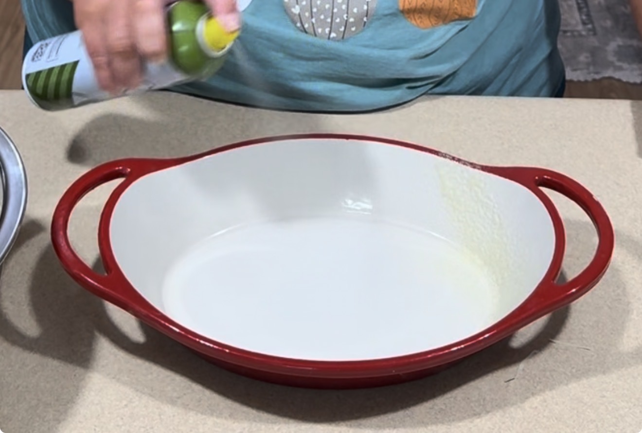 A person sprays nonstick cooking spray into a white oval baking dish with red handles, which is placed on a beige countertop.