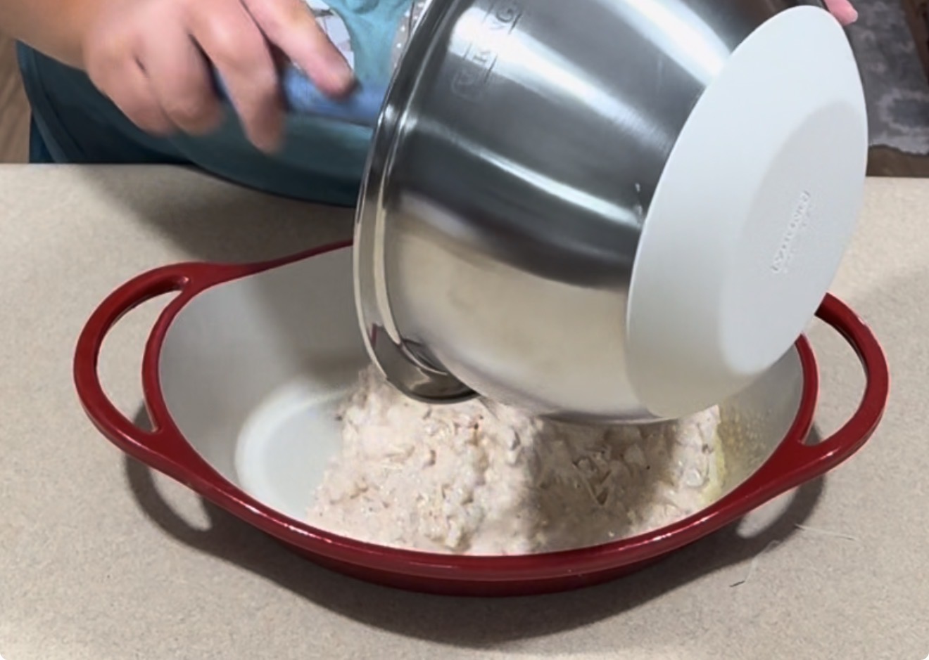 A person pours a mixture from a large metal mixing bowl into a red, oval-shaped baking dish on a countertop.
