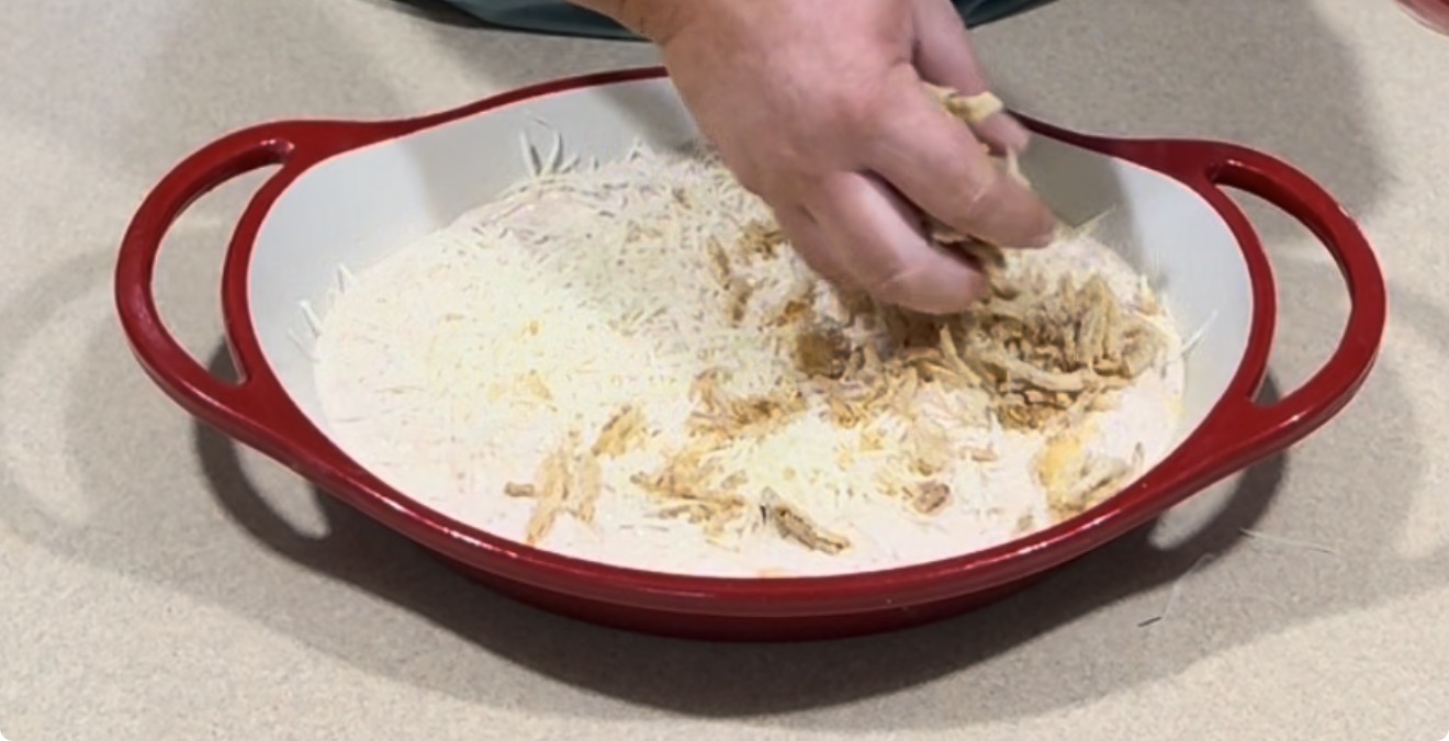 A hand is spreading shredded chicken and cheese in a red and white baking dish on a countertop, preparing ingredients for a meal.