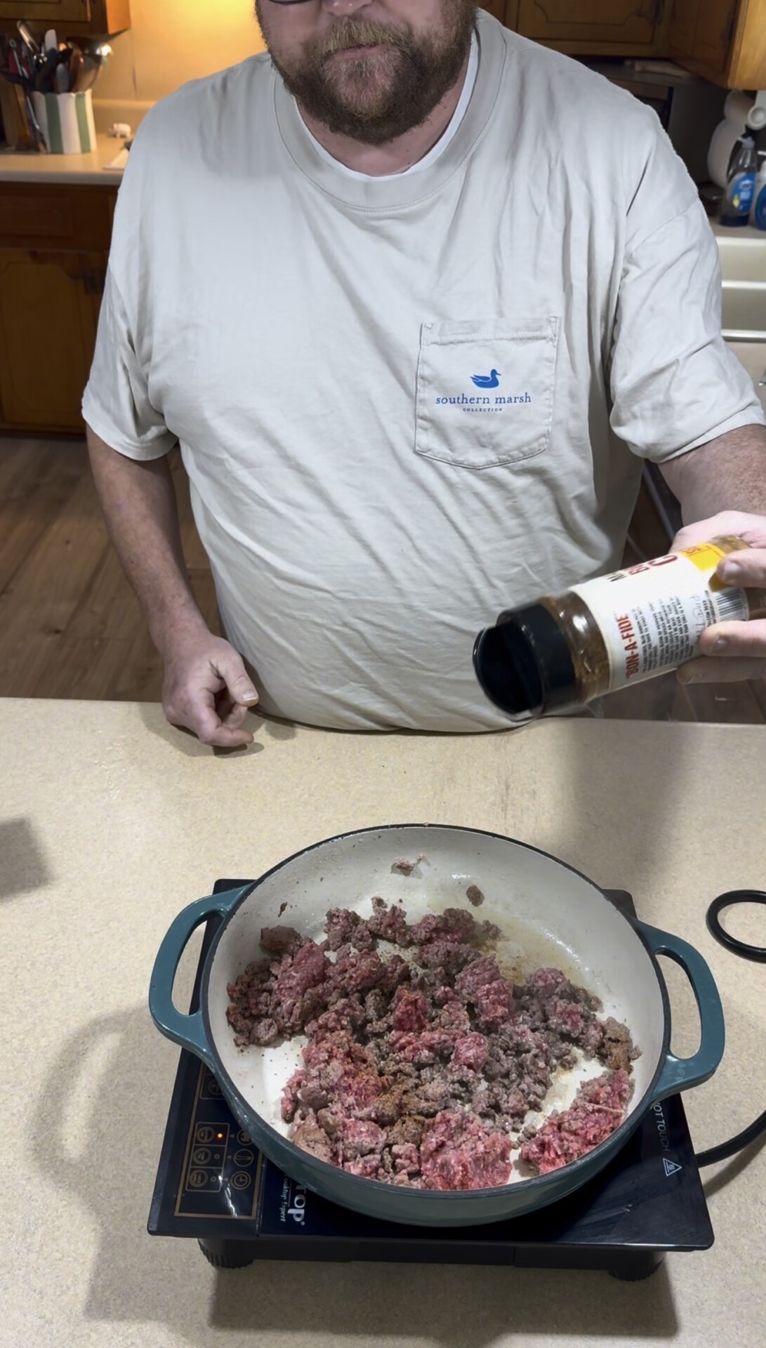 A person sprinkles seasoning from a bottle onto ground beef cooking in a skillet on a kitchen counter.