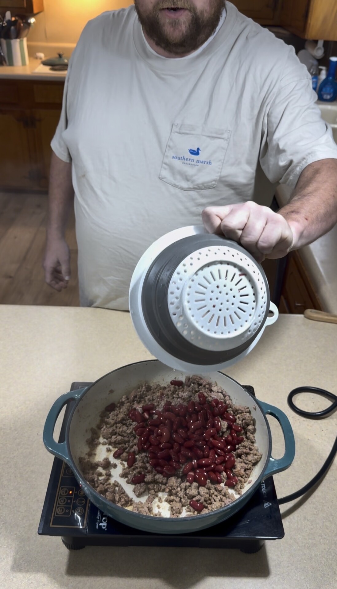 A person pours kidney beans from a colander into a pot with cooked ground beef on a stovetop in a kitchen.