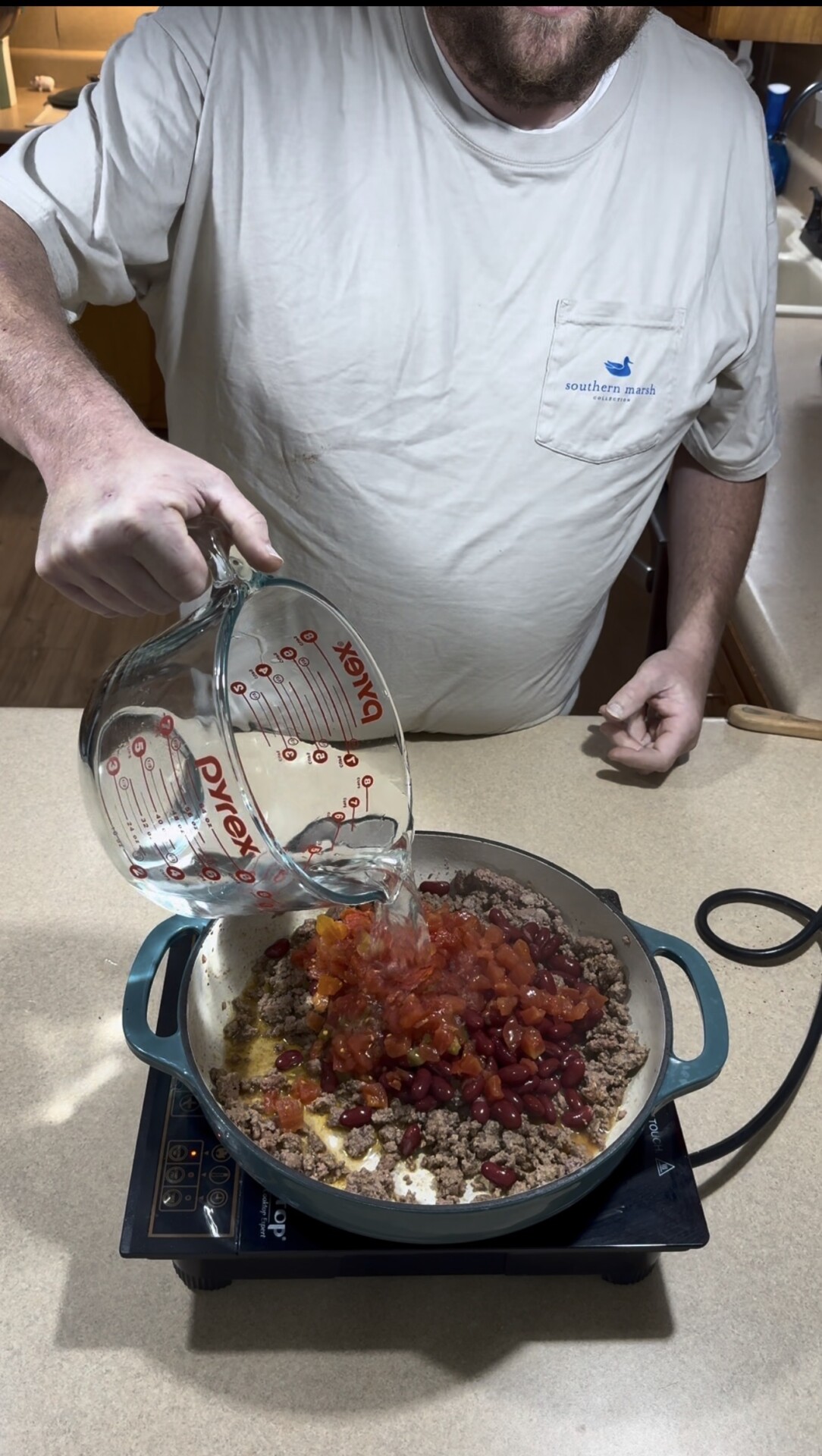 A person pours water from a Pyrex measuring cup into a skillet with ground beef, beans, diced tomatoes, and onions on a stovetop.