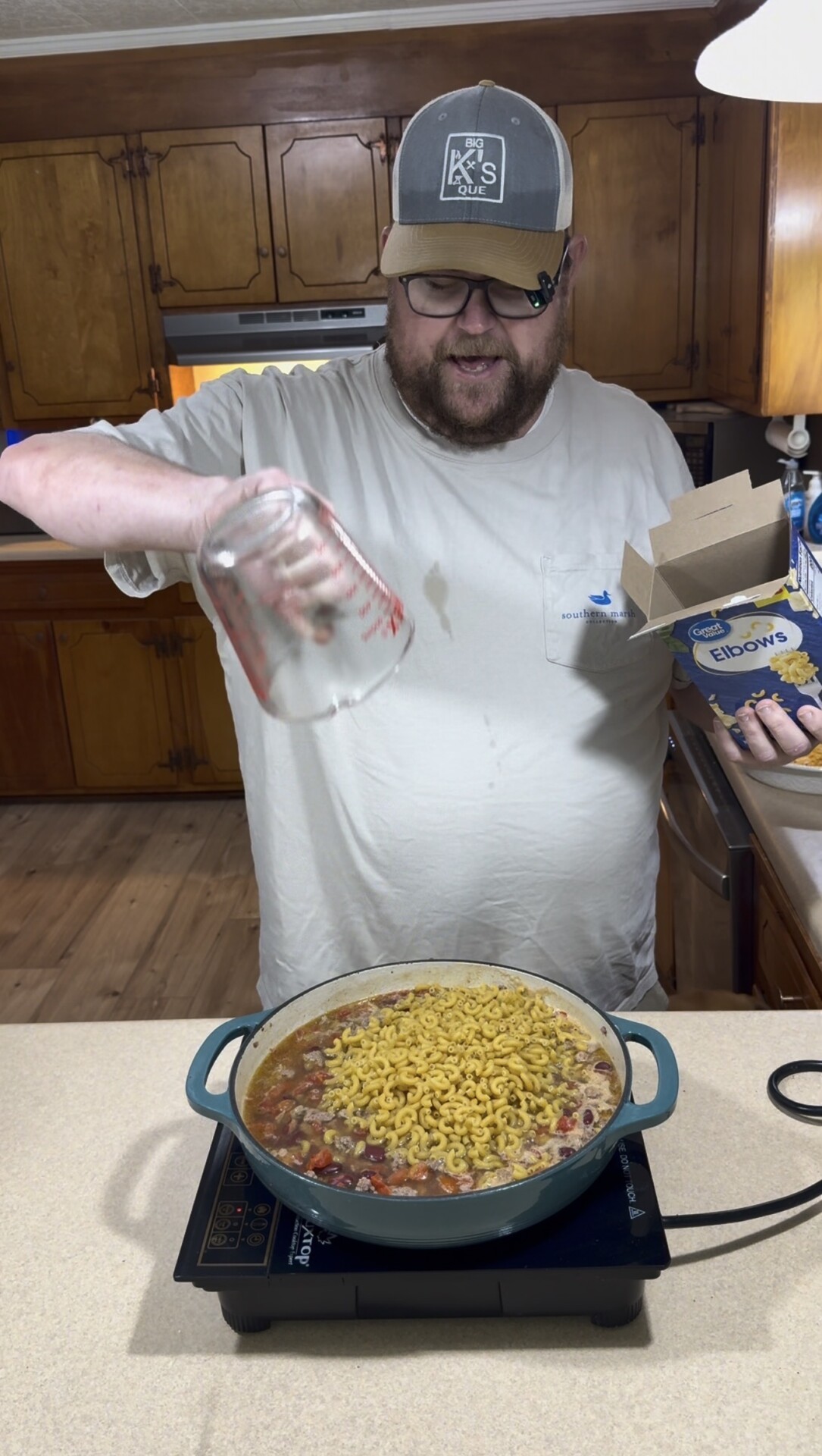 A man in a white shirt pours water from a measuring cup into a pot of soup with dry elbow macaroni on top, while holding a box of pasta in a kitchen.