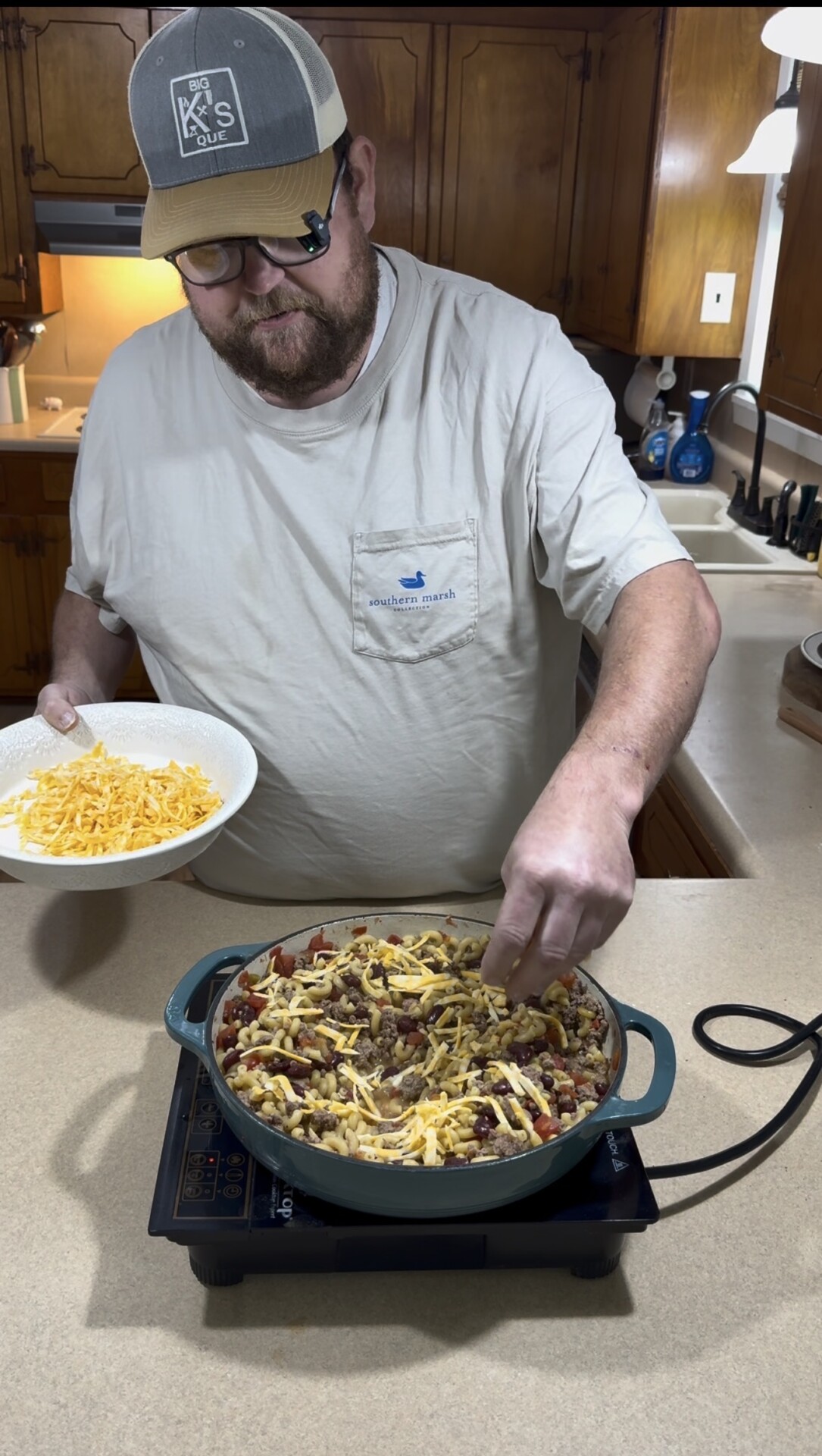 A man in a cap and glasses sprinkles shredded cheese from a bowl onto a skillet of food on a kitchen counter.