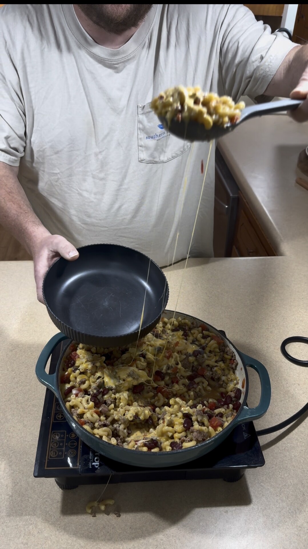A person serves cheesy pasta with beans and vegetables from a pot into a black bowl, with melted cheese stretching between the spoon and the pot on a kitchen counter.