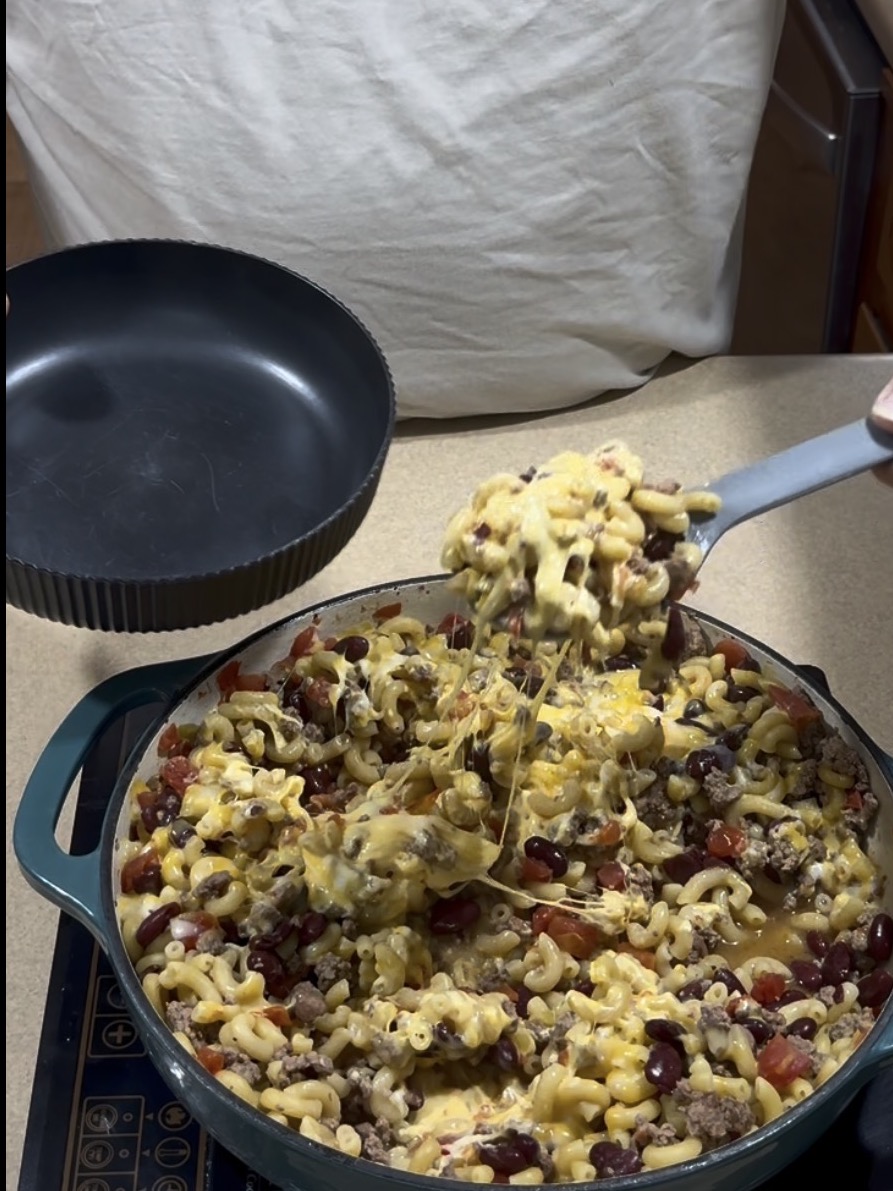A skillet filled with cheesy macaroni, ground beef, beans, and tomatoes is being served with a spoon, with some cheese stretching from the pan.