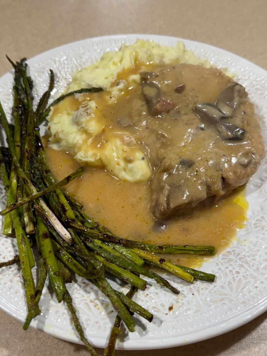 A plate with mashed potatoes topped with brown mushroom gravy, a slice of meat, and a side of roasted asparagus spears on a white patterned dish.