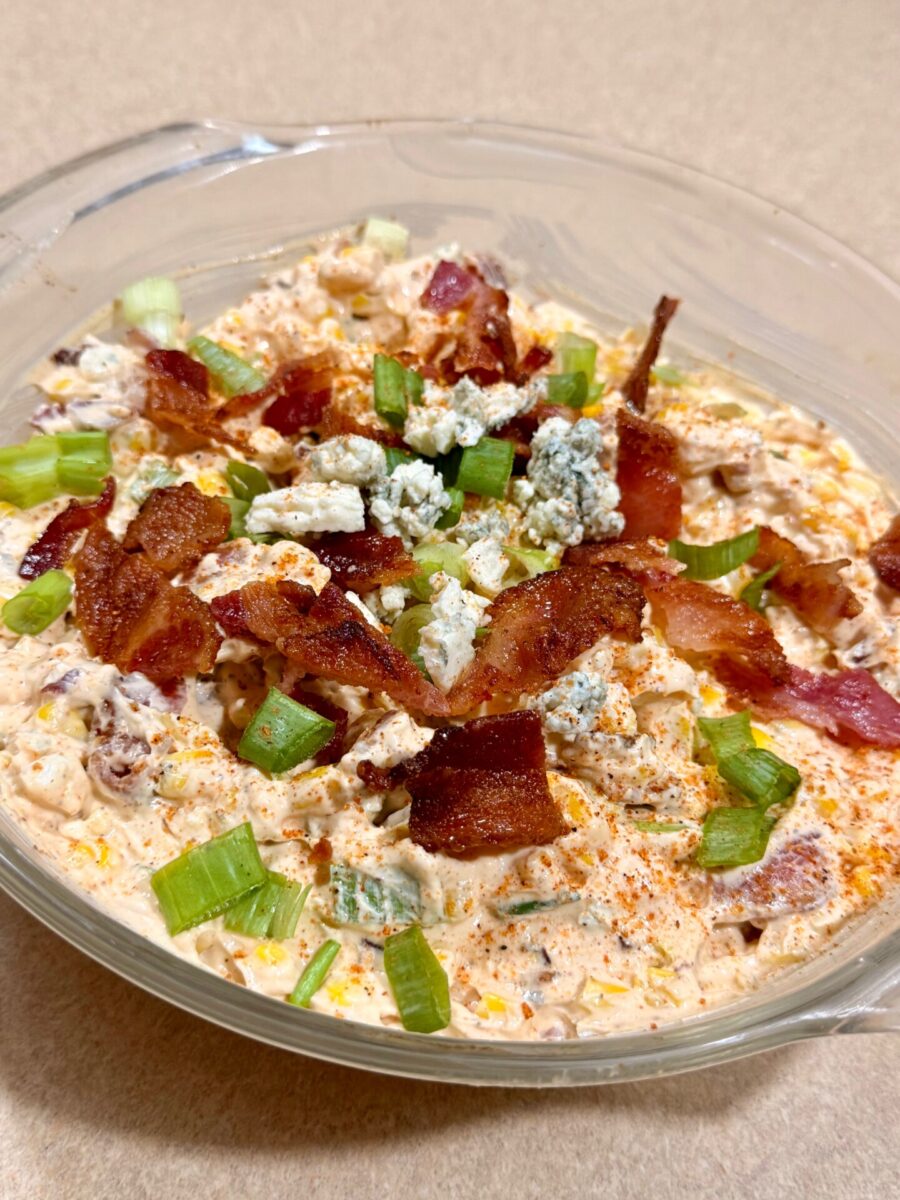 Glass bowl filled with creamy dip, topped with crumbled bacon, blue cheese, chopped green onions, and a sprinkle of paprika, on a beige counter.