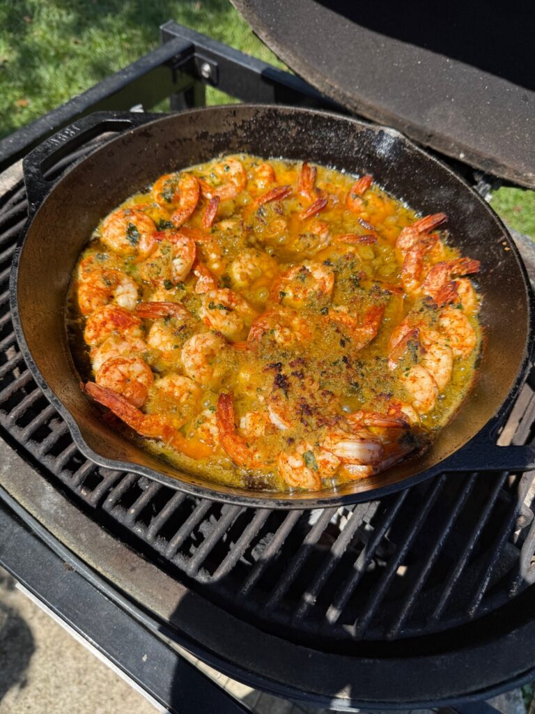 A cast iron skillet filled with shrimp cooking in a seasoned, yellow-orange sauce sits on a grill outdoors, with sunlight highlighting the dish.