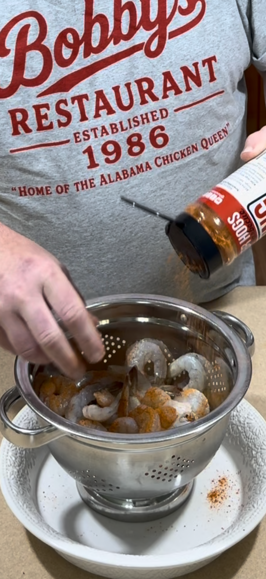 A person sprinkles seasoning onto raw shrimp in a metal colander, wearing a gray Bobbys Restaurant shirt. The colander sits on a white plate on a counter.