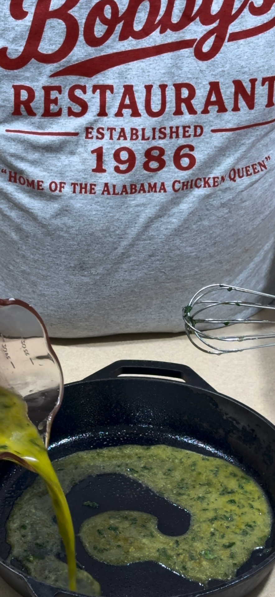 Egg mixture is being poured into a cast iron skillet as a whisk rests nearby. In the background, a shirt reads, Bobbys Restaurant Established 1986 Home of the Alabama Chicken Queen..