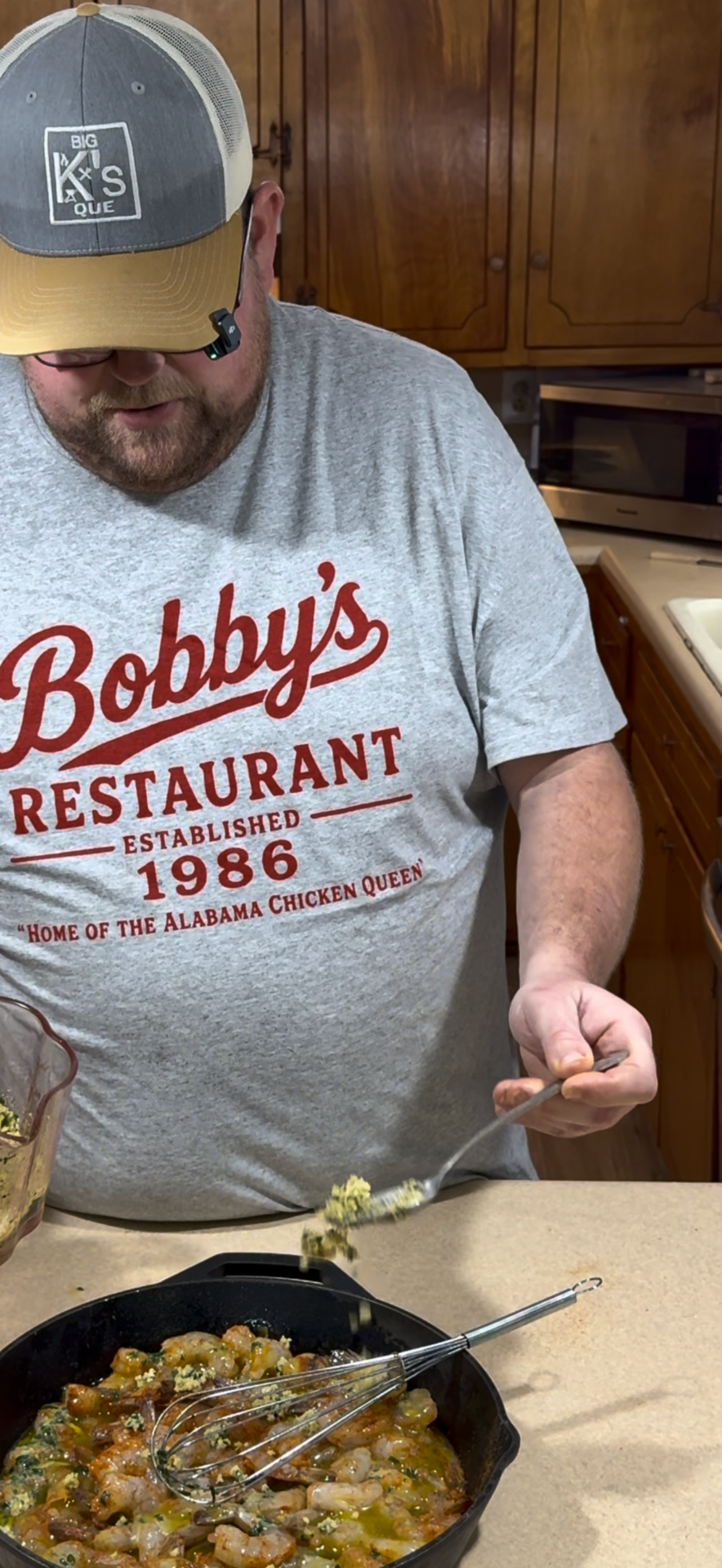A man in a Bobbys Restaurant shirt spoons food from a glass dish onto a plate in a kitchen with wooden cabinets.