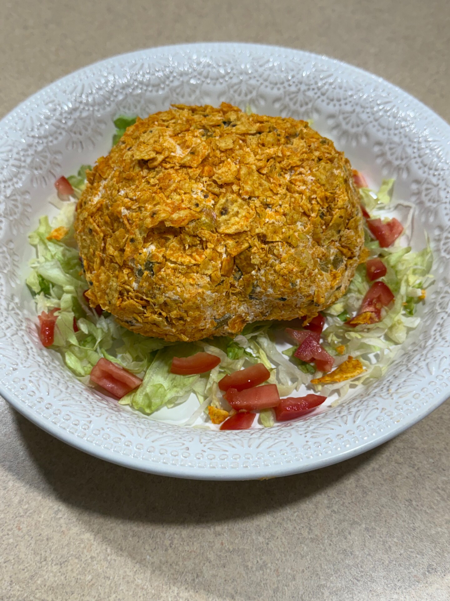 A round, orange-crusted ball sits on chopped lettuce and diced tomatoes in a white textured bowl, placed on a tan countertop.
