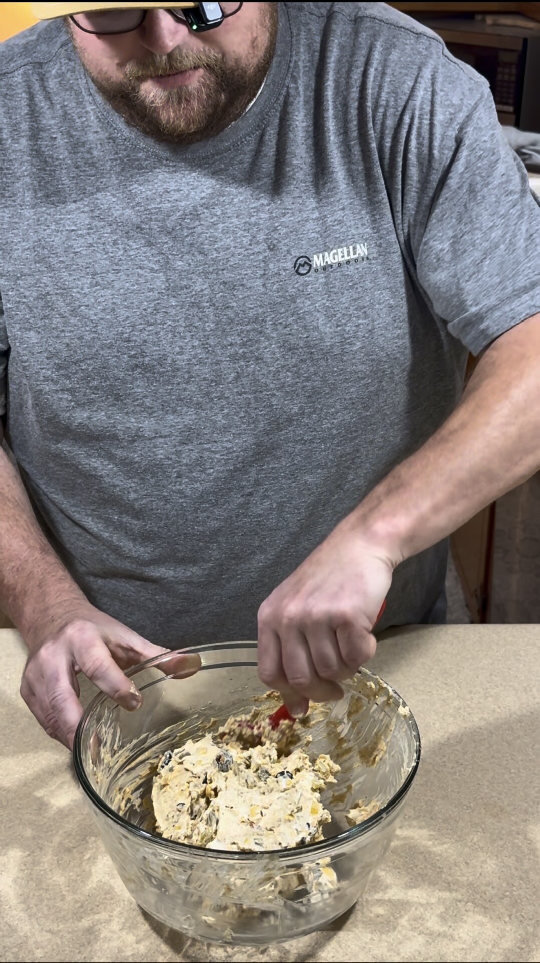A person in a gray shirt mixes cookie dough with a spatula in a glass bowl on a beige countertop.