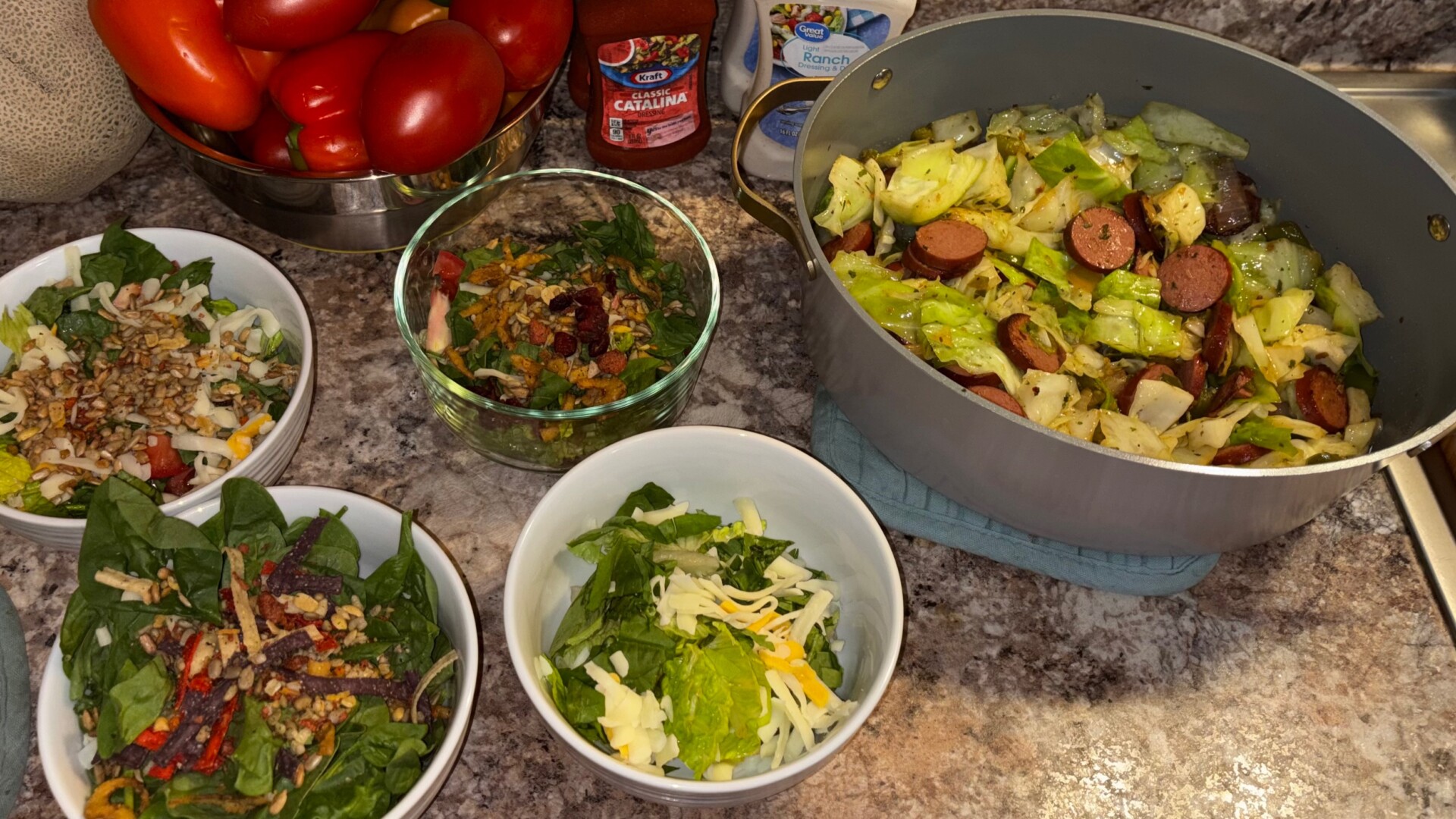 Bowls of salad with toppings, shredded cheese, a large pot of cabbage and sausage, tomatoes in a bowl, dressing packets, and a cantaloupe on a kitchen counter.