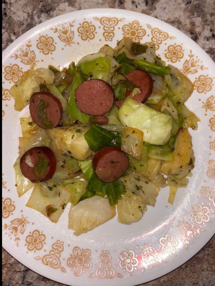 A plate of cooked cabbage mixed with sliced sausage, garnished with herbs, served on a white plate with a gold floral pattern.