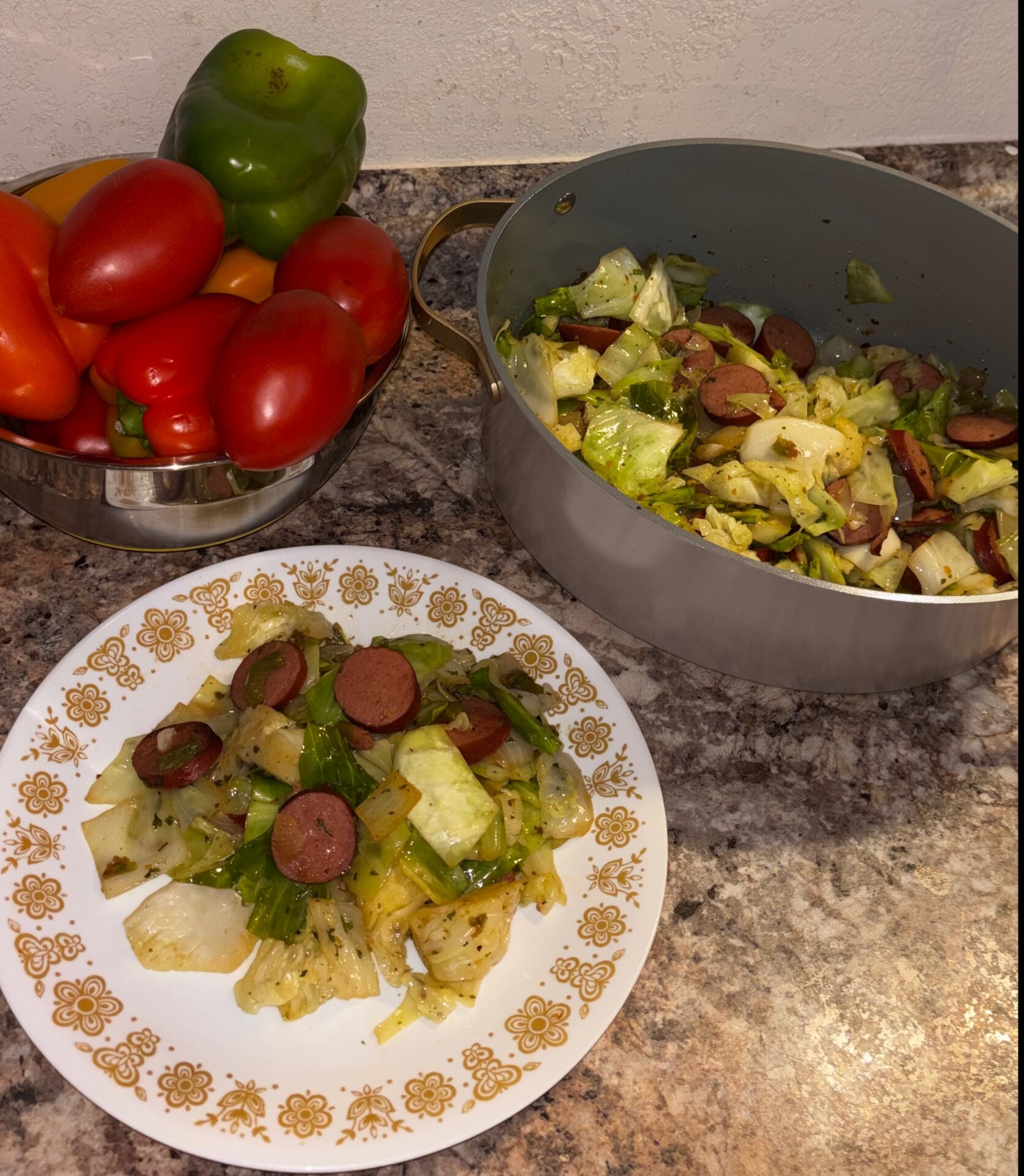 A plate of cooked cabbage and sausage sits beside a pot of the same dish. Nearby, a bowl holds fresh bell peppers and tomatoes on a marbled kitchen counter.