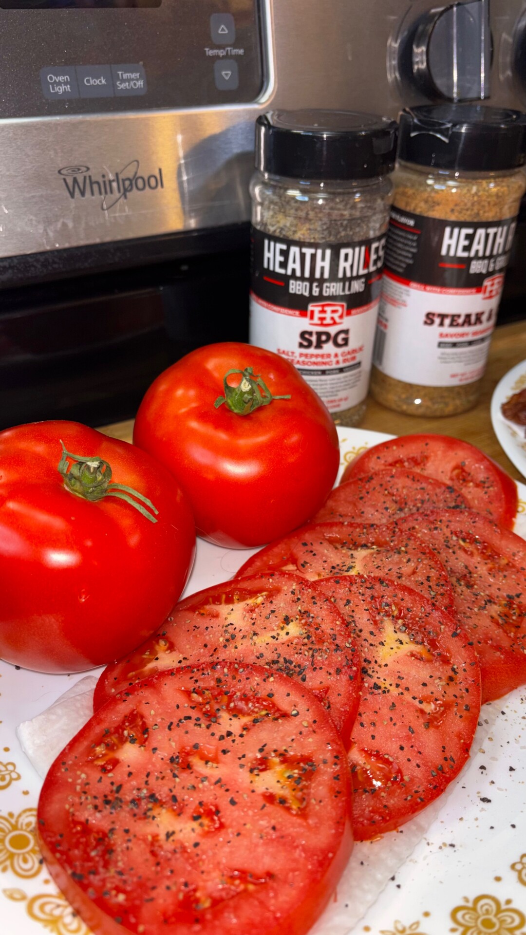 Two whole tomatoes and several tomato slices sprinkled with spices on a plate. Behind them are two bottles of Heath Riles seasoning and part of a Whirlpool appliance.
