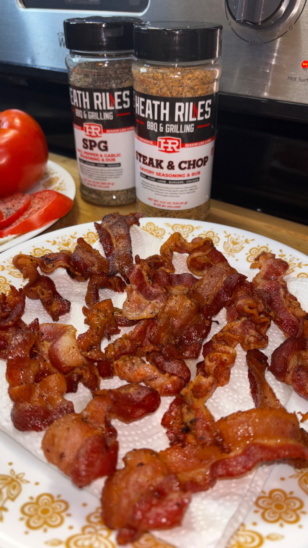 A plate of crispy cooked bacon sits on a paper towel. Behind it are Heath Riles BBQ & Grilling SPG and Steak & Chop seasoning bottles, with a sliced tomato beside them on a kitchen countertop.