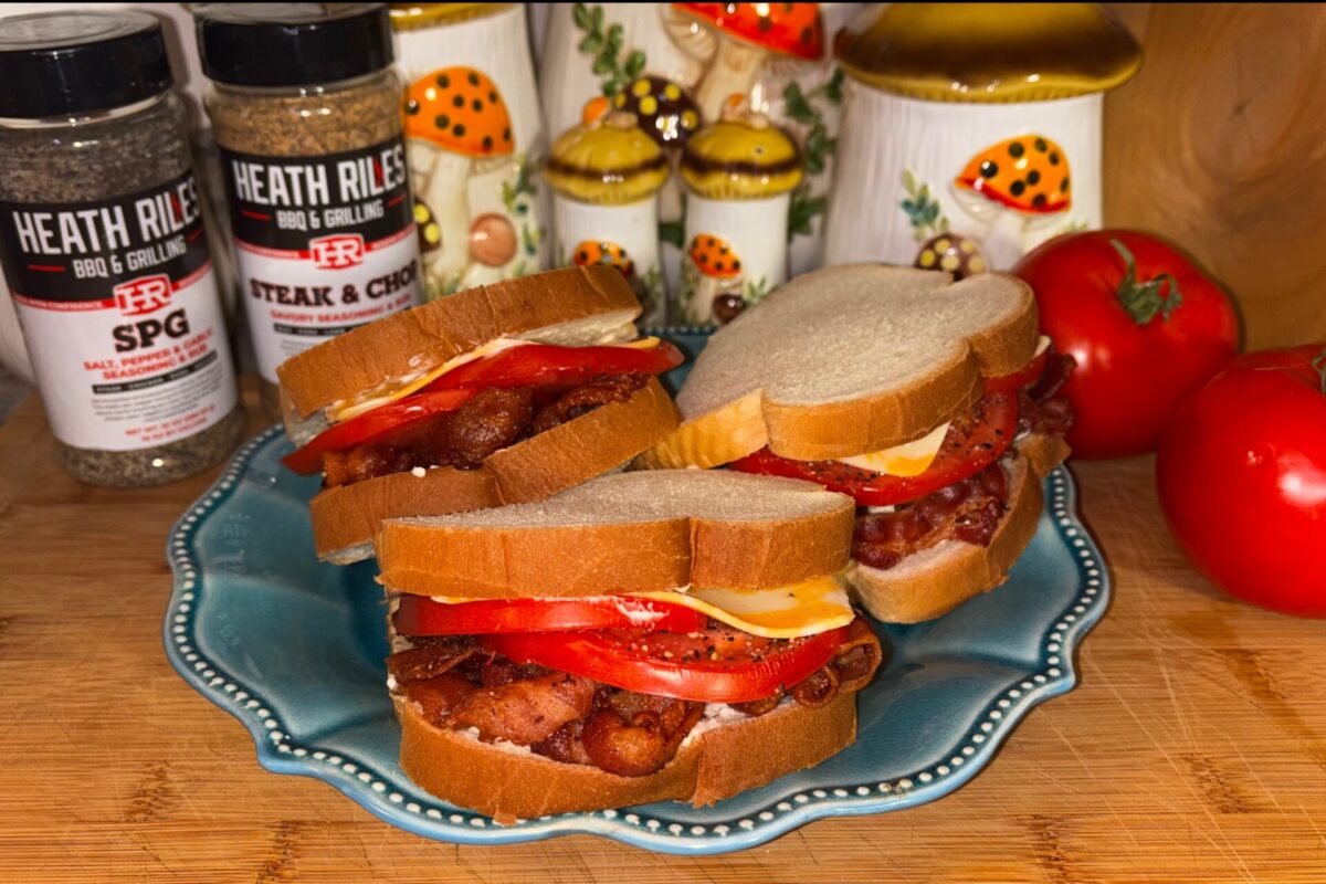 Three BLT sandwiches with bacon, lettuce, tomato, and cheese on a blue plate, with tomatoes, spice jars, and decorative mushroom canisters in the background.