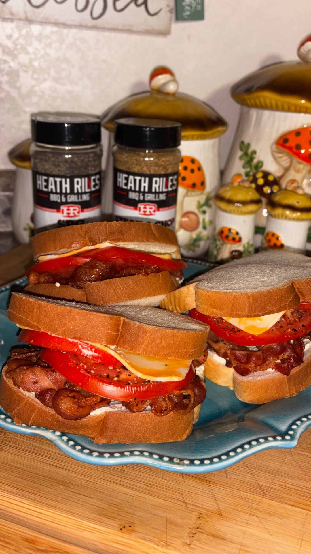 Three BLT sandwiches with bacon, tomato, cheese, and mayonnaise on white bread are arranged on a blue plate. Two seasoning containers and ceramic mushroom-themed canisters are in the background.