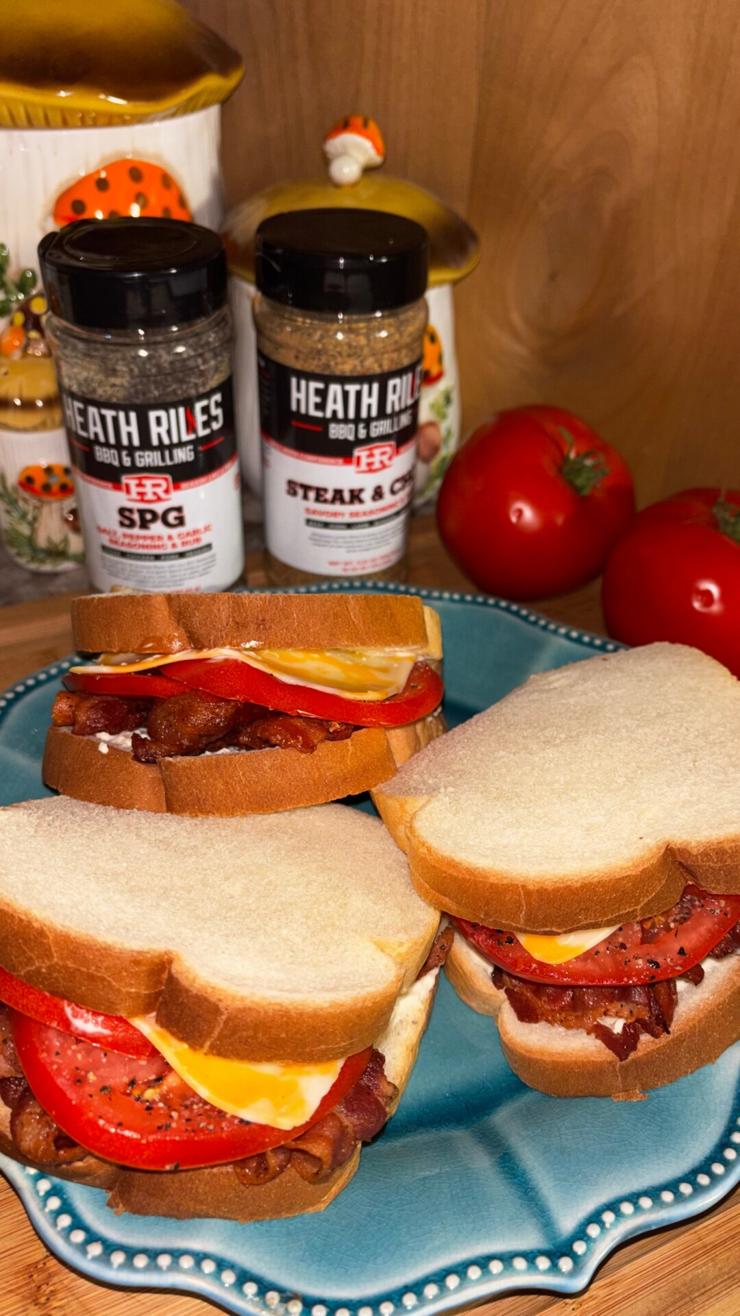 Three BLT sandwiches on a blue plate, each with bacon, tomato, and cheese on white bread. Two tomatoes and two Heath Riles seasoning bottles are in the background on a wooden surface.