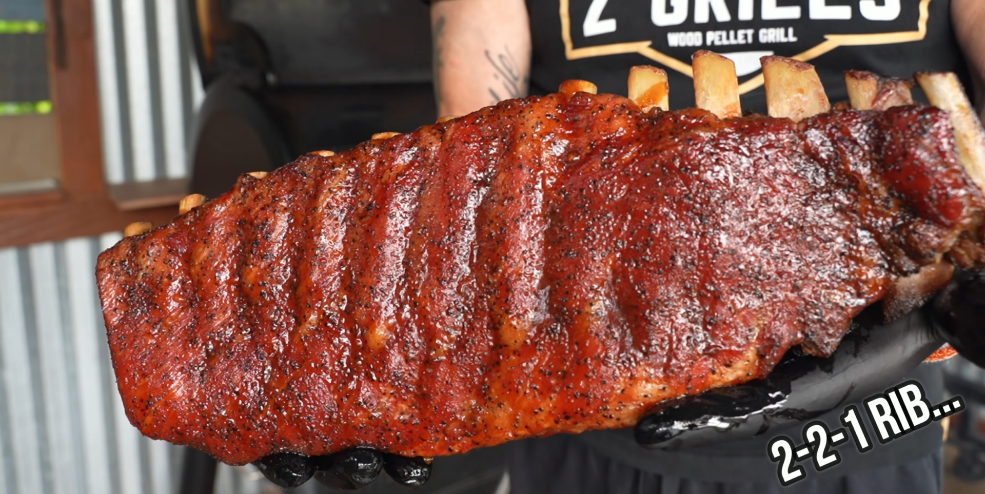 A person wearing black gloves holds a large, cooked rack of ribs with a dark seasoned crust. The ribs are presented close to the camera. 2-2-1 RIB… is written in the lower right corner.