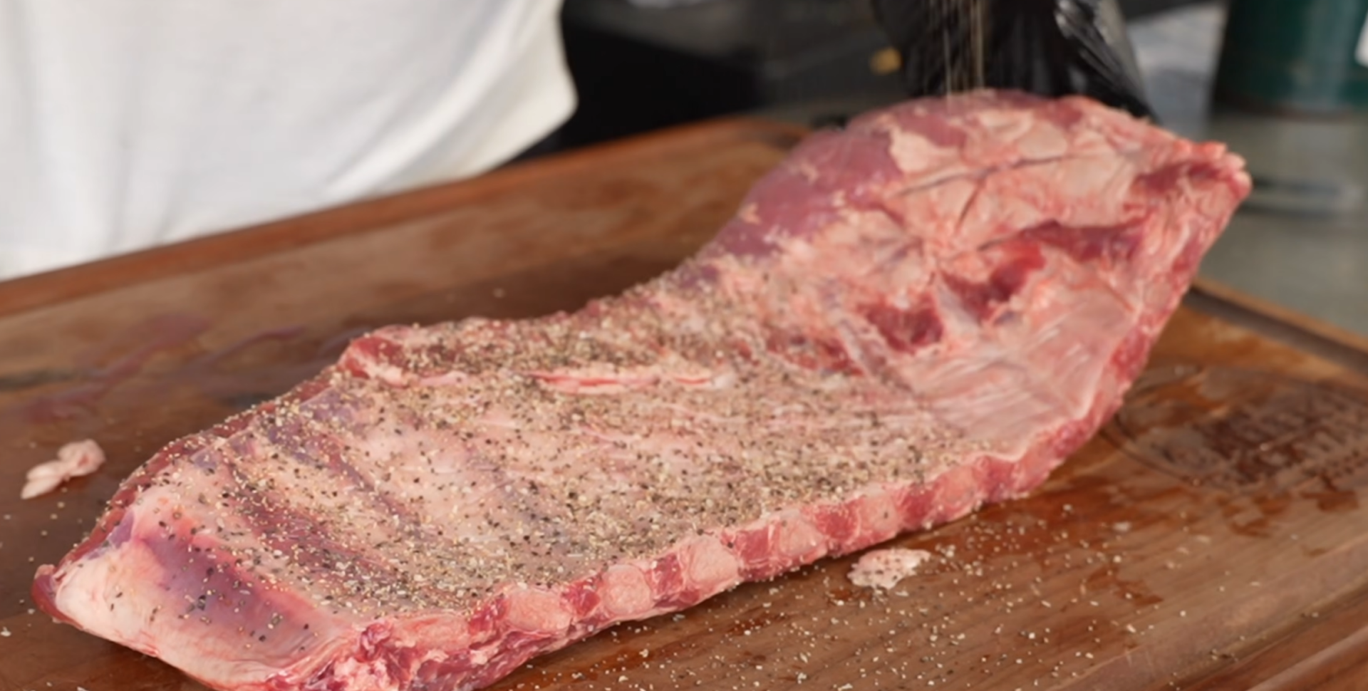A large raw rack of ribs is seasoned with salt and pepper on a wooden cutting board. A person is sprinkling seasoning over the meat, wearing a white shirt and a black glove.