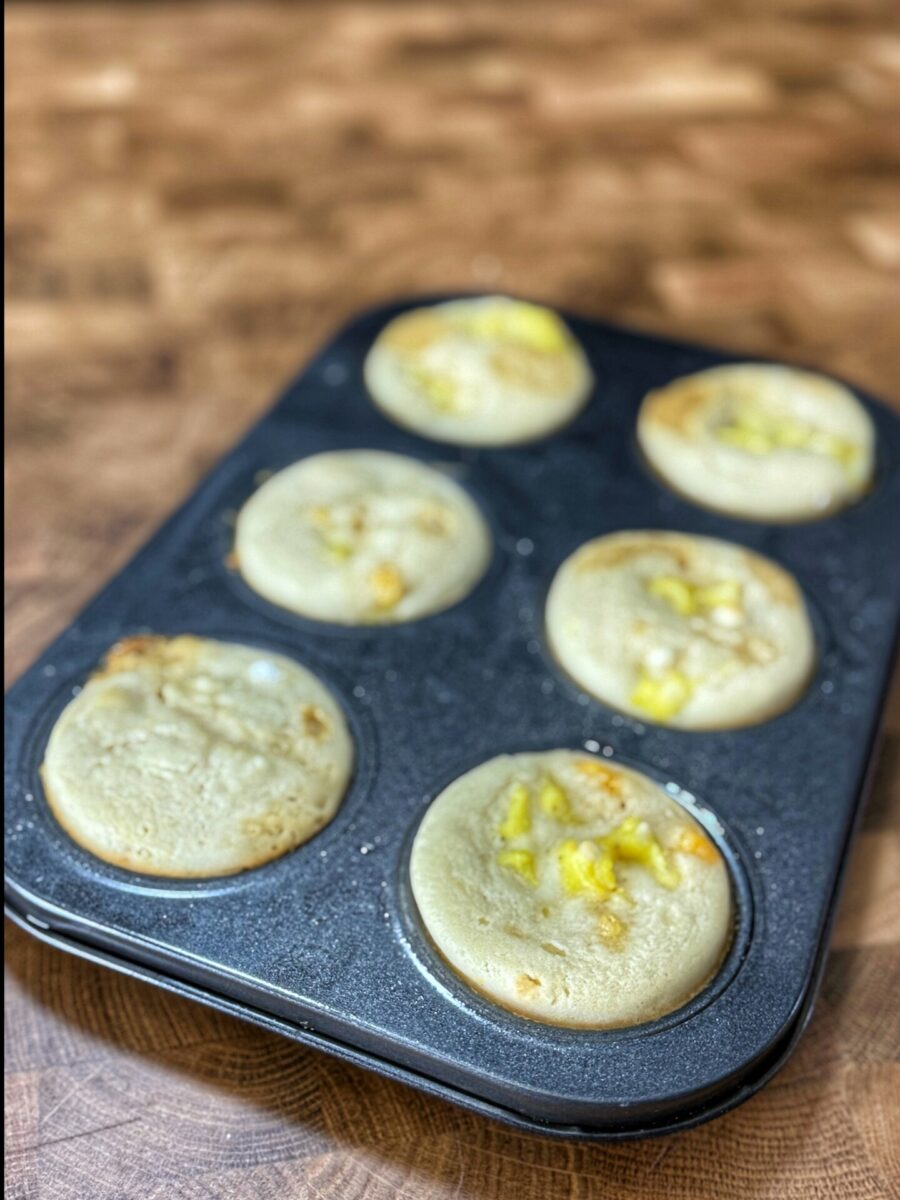 A muffin tin holds seven round, pale pancakes with bits of yellow fruit or cheese on top, resting on a wooden surface.