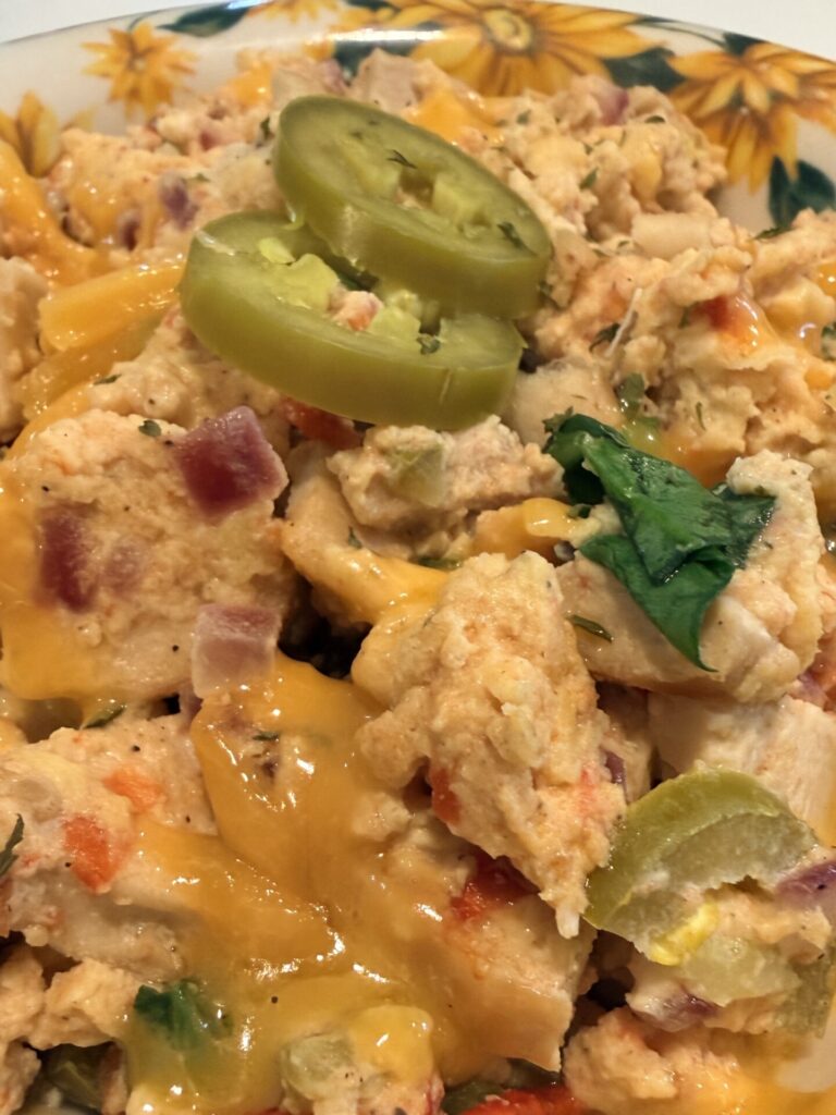 Scrambled eggs with melted cheddar cheese, diced vegetables, and herbs, topped with two slices of pickled jalapeño, served on a floral-patterned plate.