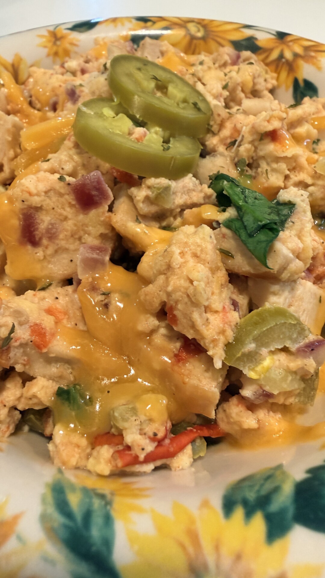 Scrambled eggs with melted cheddar cheese, chopped red onions, tomatoes, spinach, and topped with sliced jalapeños, served in a bowl with a sunflower pattern.