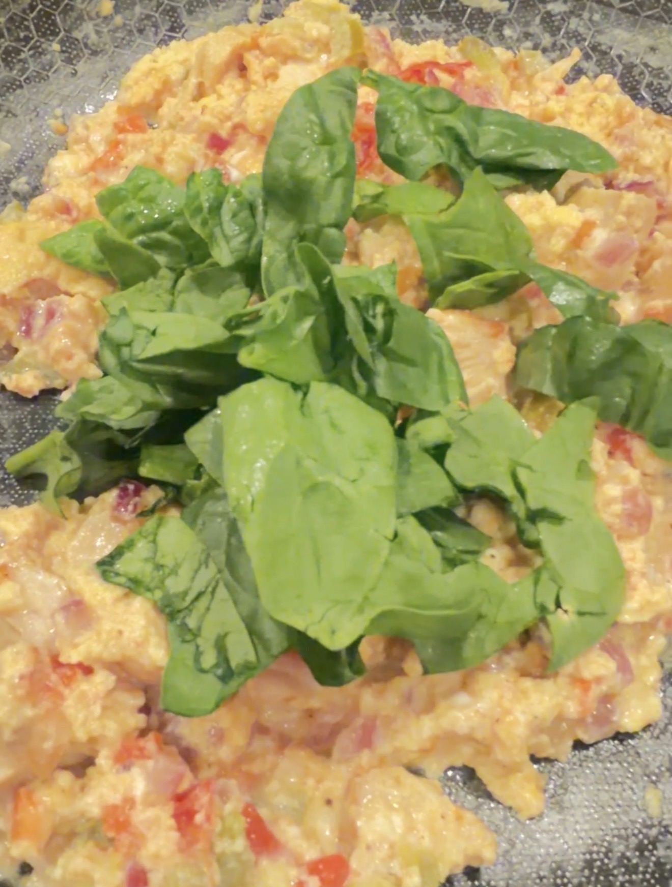 Scrambled eggs with chopped tomatoes and onions, topped with fresh spinach leaves, on a dark plate.
