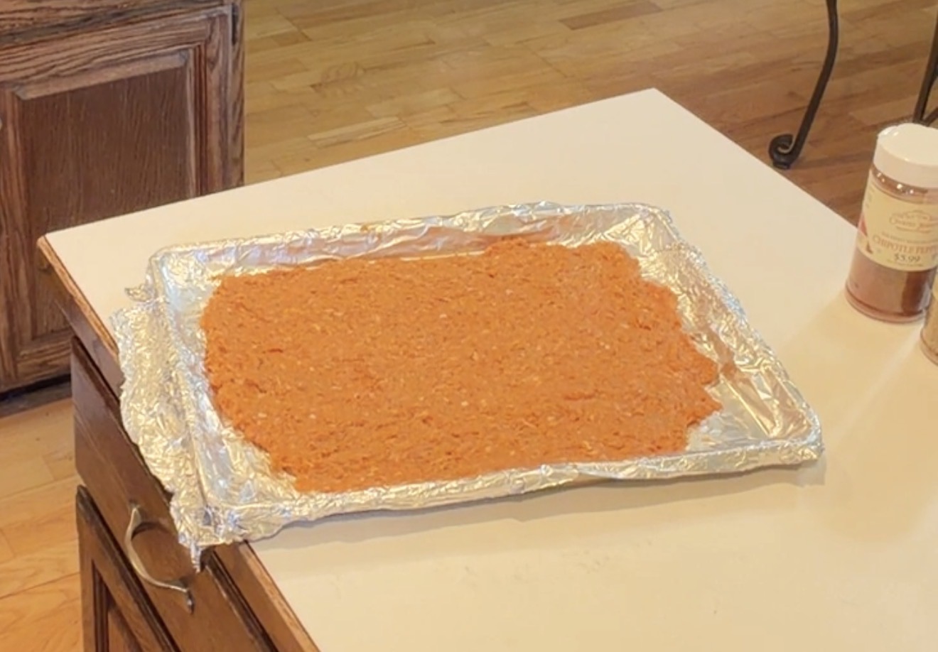 A baking sheet lined with foil holding a layer of spread, orange-colored ground meat mixture, sitting on a kitchen counter next to a spice jar.