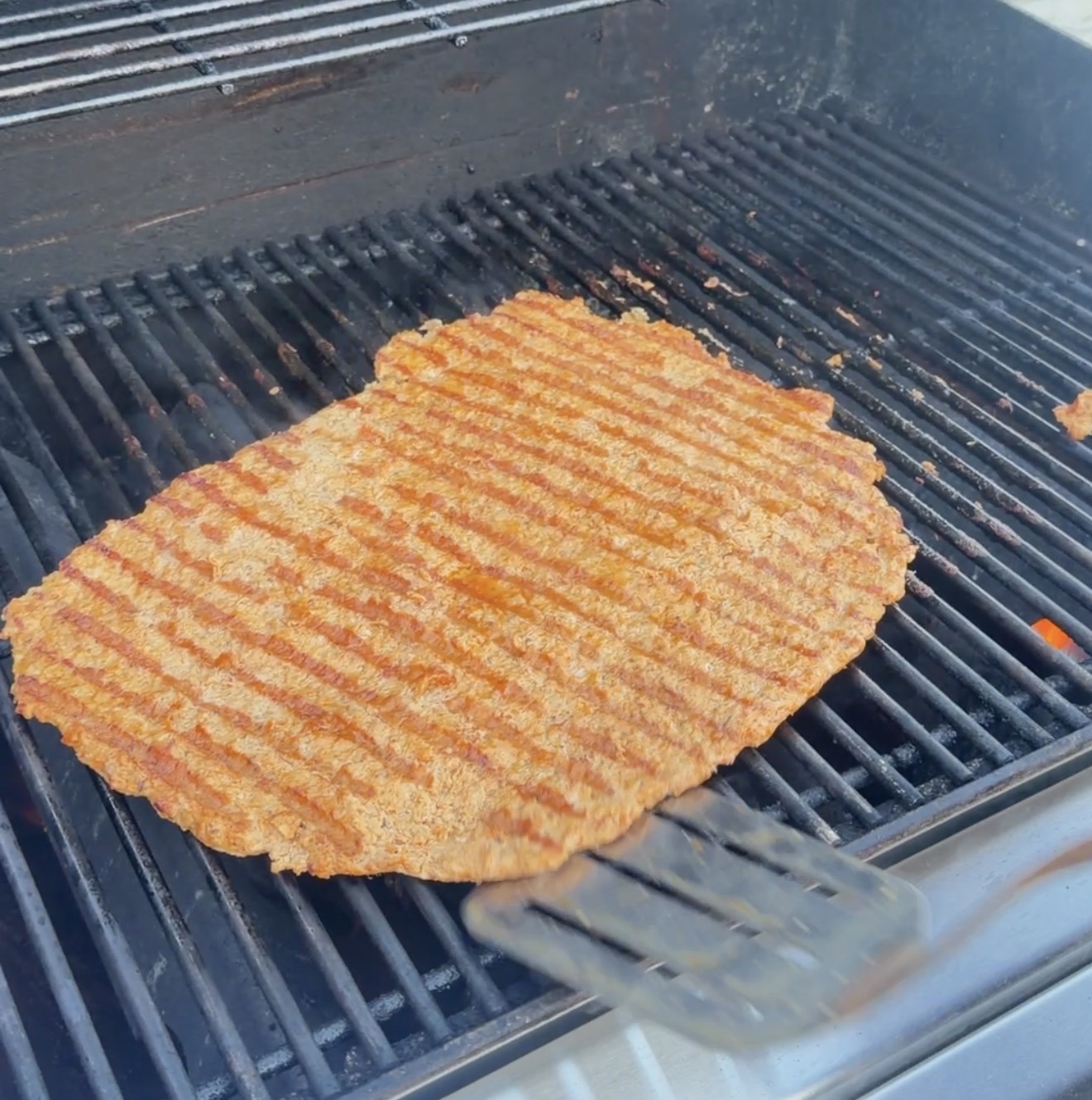 A large, thin burger patty with dark grill marks cooking on a metal barbecue grate.