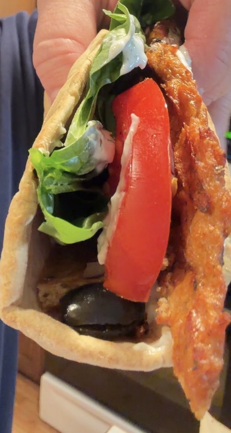 A hand holding a pita wrap filled with a breaded chicken strip, a slice of tomato, lettuce, a black olive, and sauce.