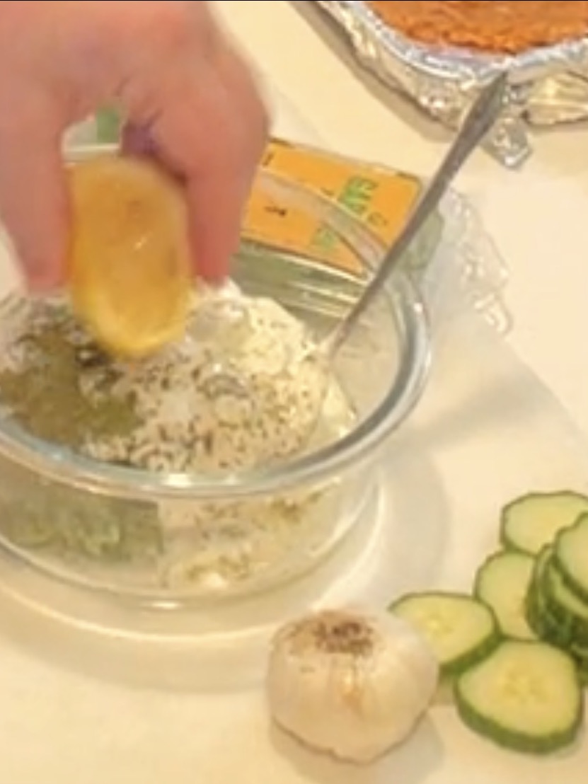A hand squeezes a lemon wedge into a glass bowl of dip topped with herbs, next to a bulb of garlic and sliced cucumbers.