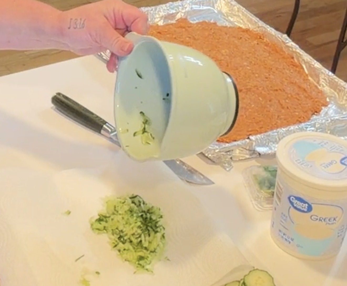 A hand pours shredded cucumber from a small green bowl onto a paper towel, near a baking sheet of ground meat and a container of Greek yogurt.