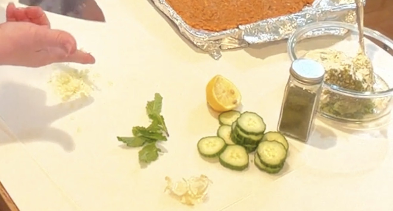A hand sprinkles minced garlic onto a counter next to fresh mint leaves, sliced cucumbers, half a lemon, a spice jar, and a bowl of dip.