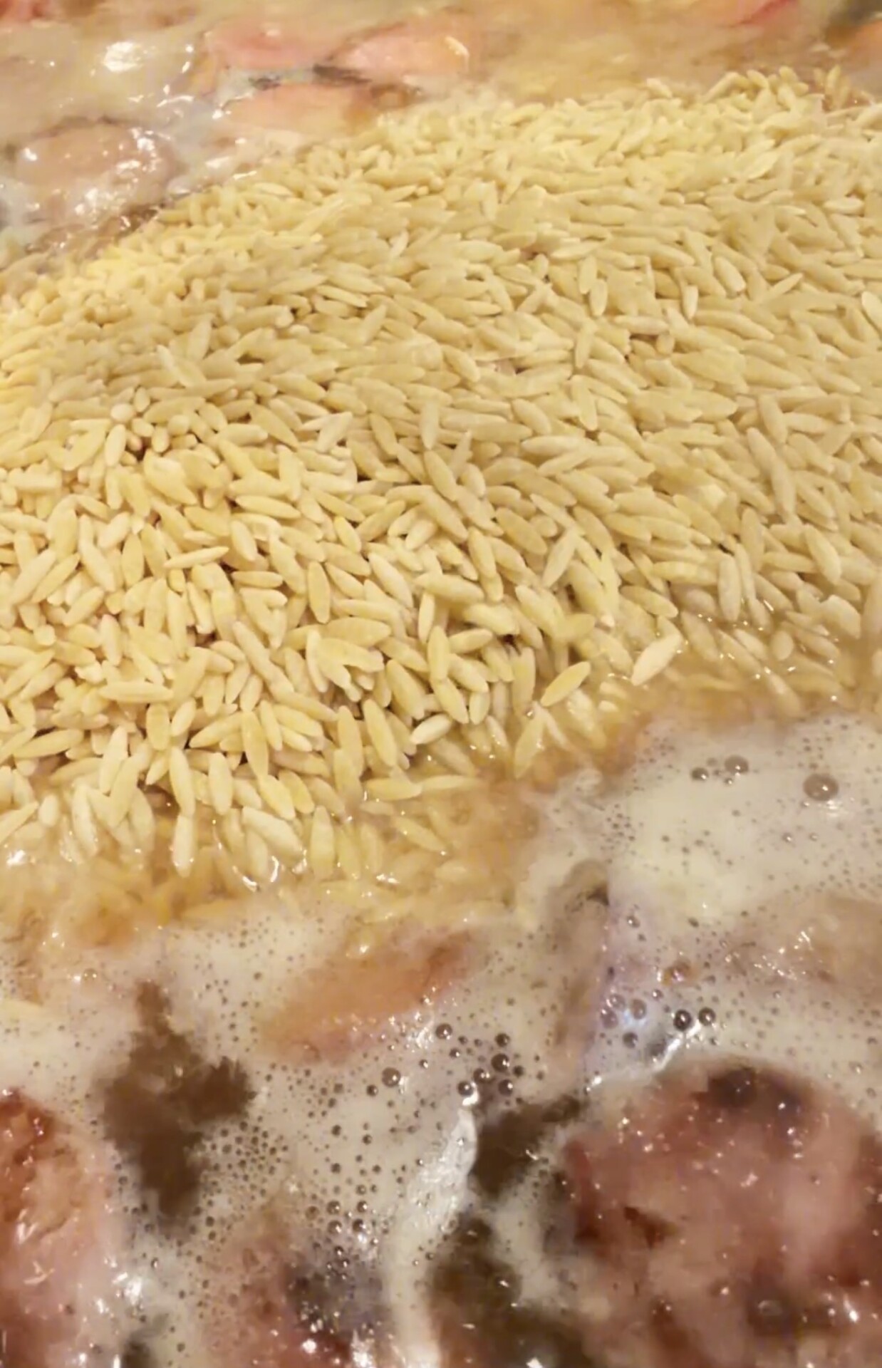 Uncooked orzo pasta floating in a pot of simmering liquid with chunks of meat.