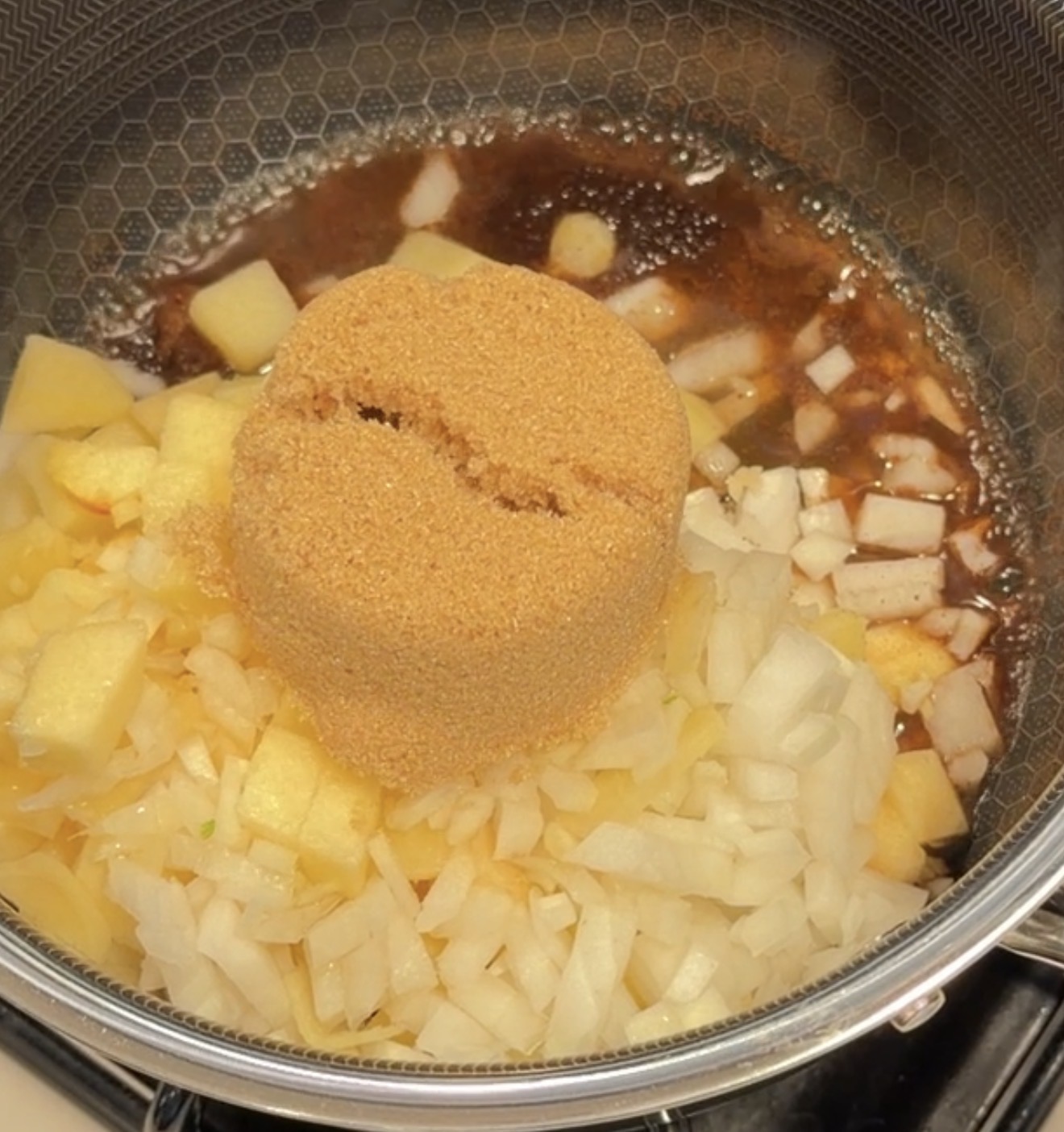 A mound of brown sugar sits atop chopped onions and diced apples in a simmering pan of sauce.