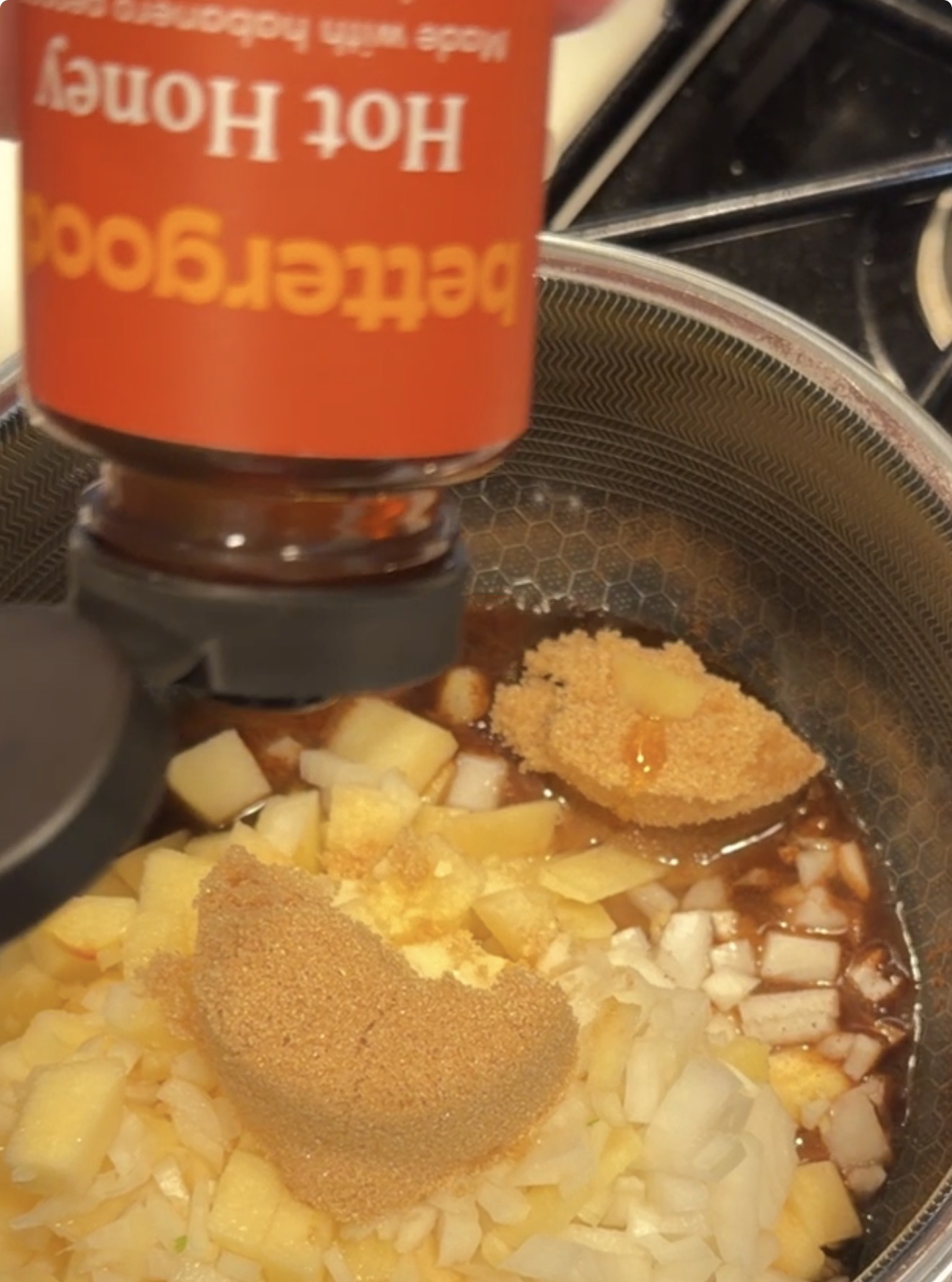 A hand squeezes a bottle of Bettergoods Hot Honey over a pot containing diced apples, chopped onions, and scoops of brown sugar.