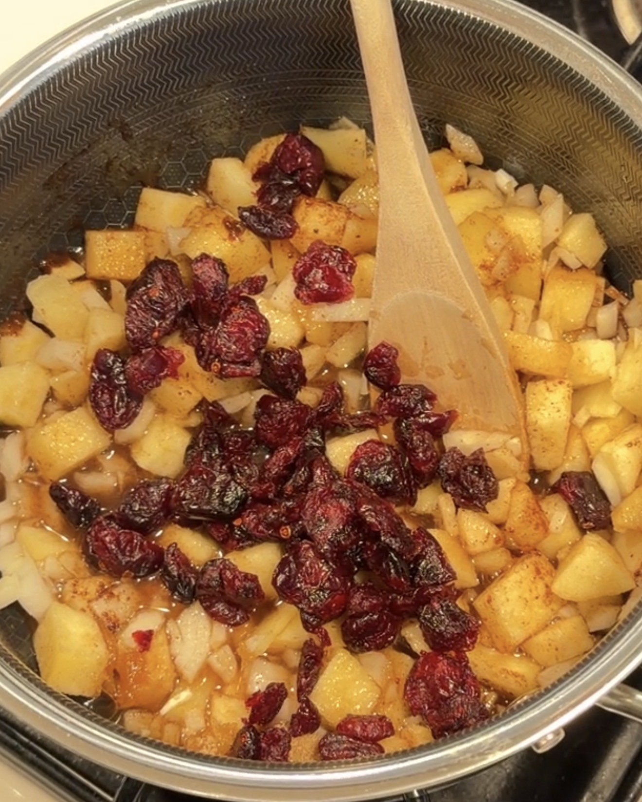A wooden spoon stirs diced apples, onions, and dried cranberries in a metal pot.