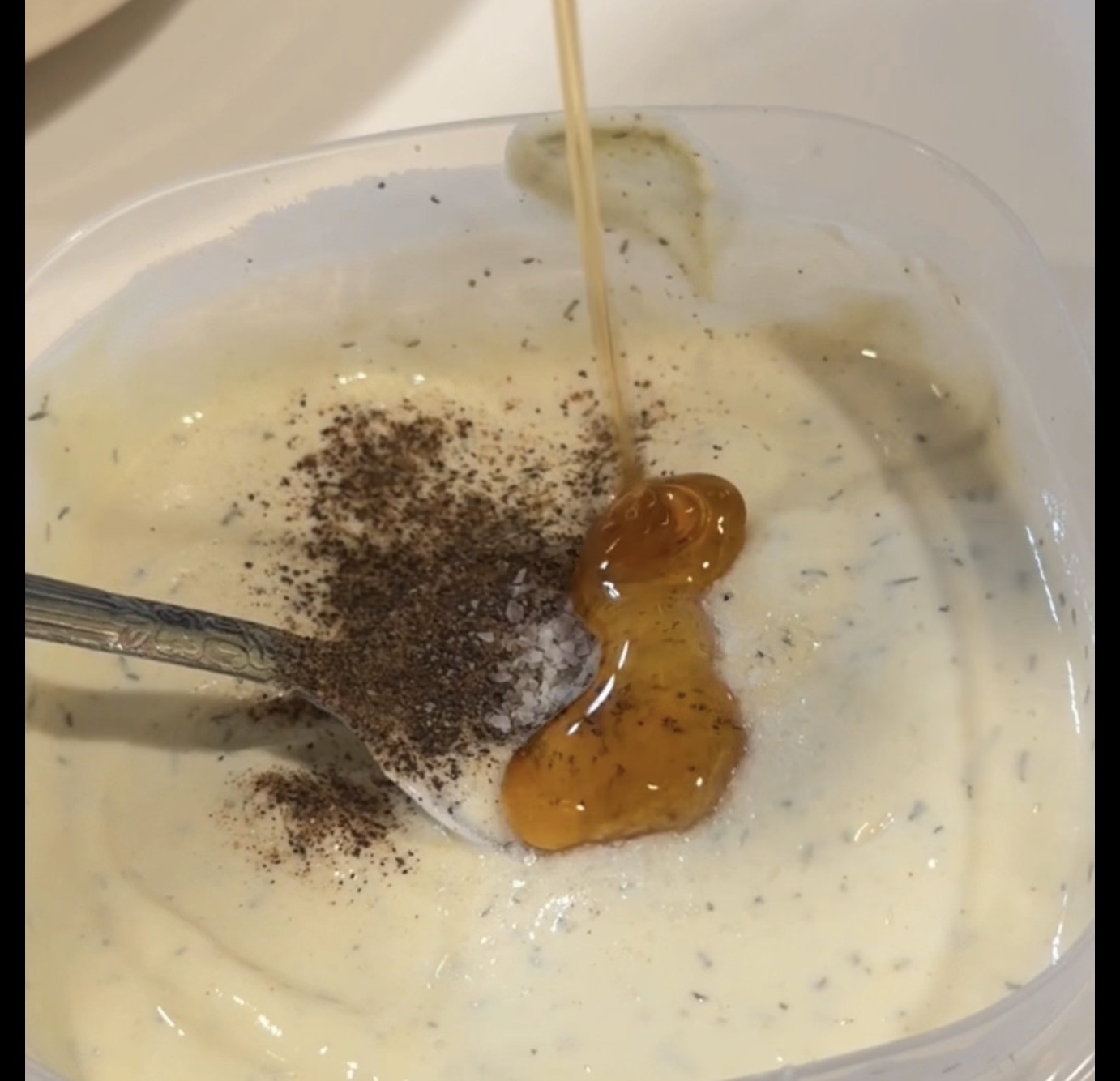 Honey being drizzled into a plastic container of creamy herb dressing, with a spoonful of black pepper resting in the mixture.