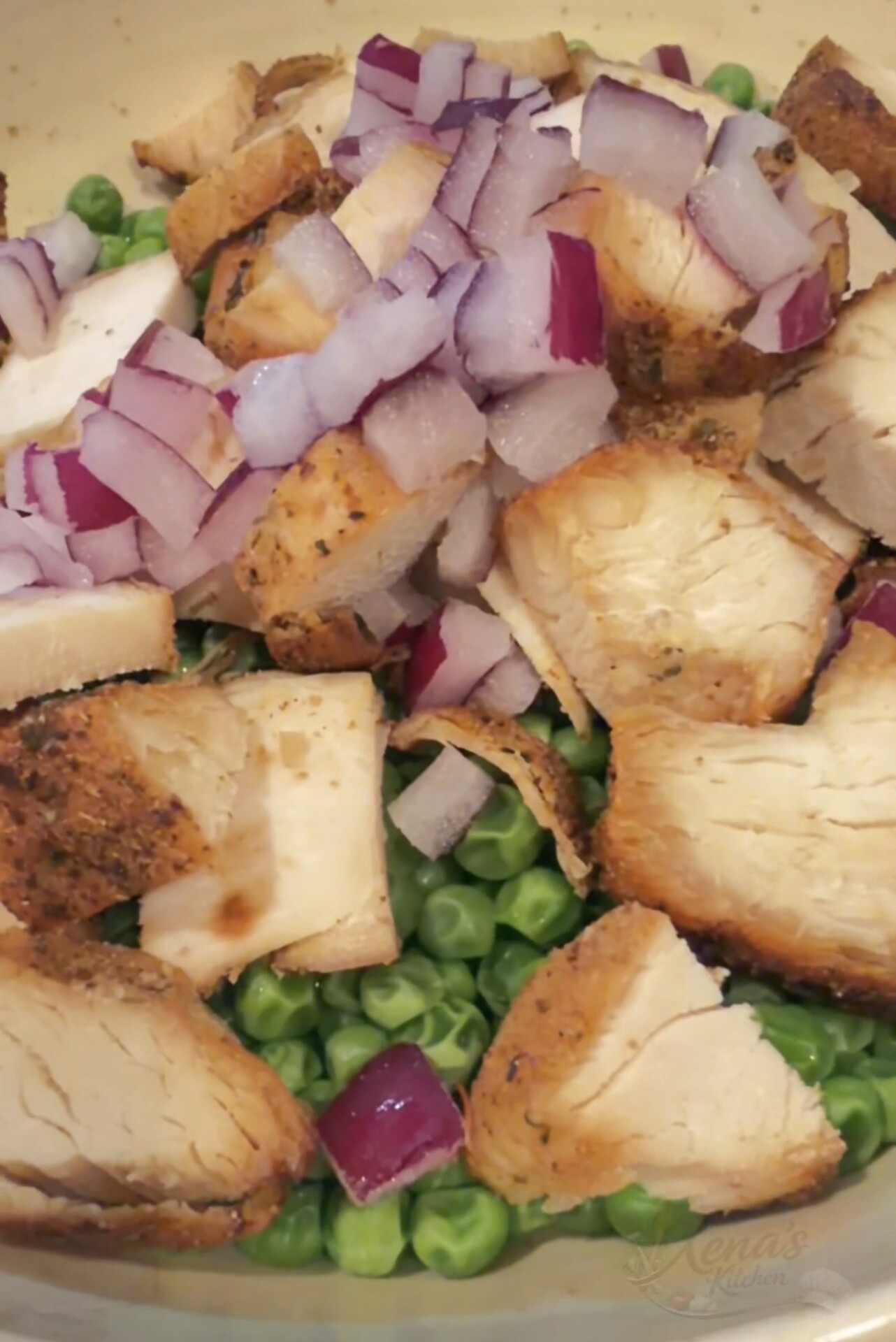 A bowl of green peas topped with pieces of seasoned chicken and diced red onion.